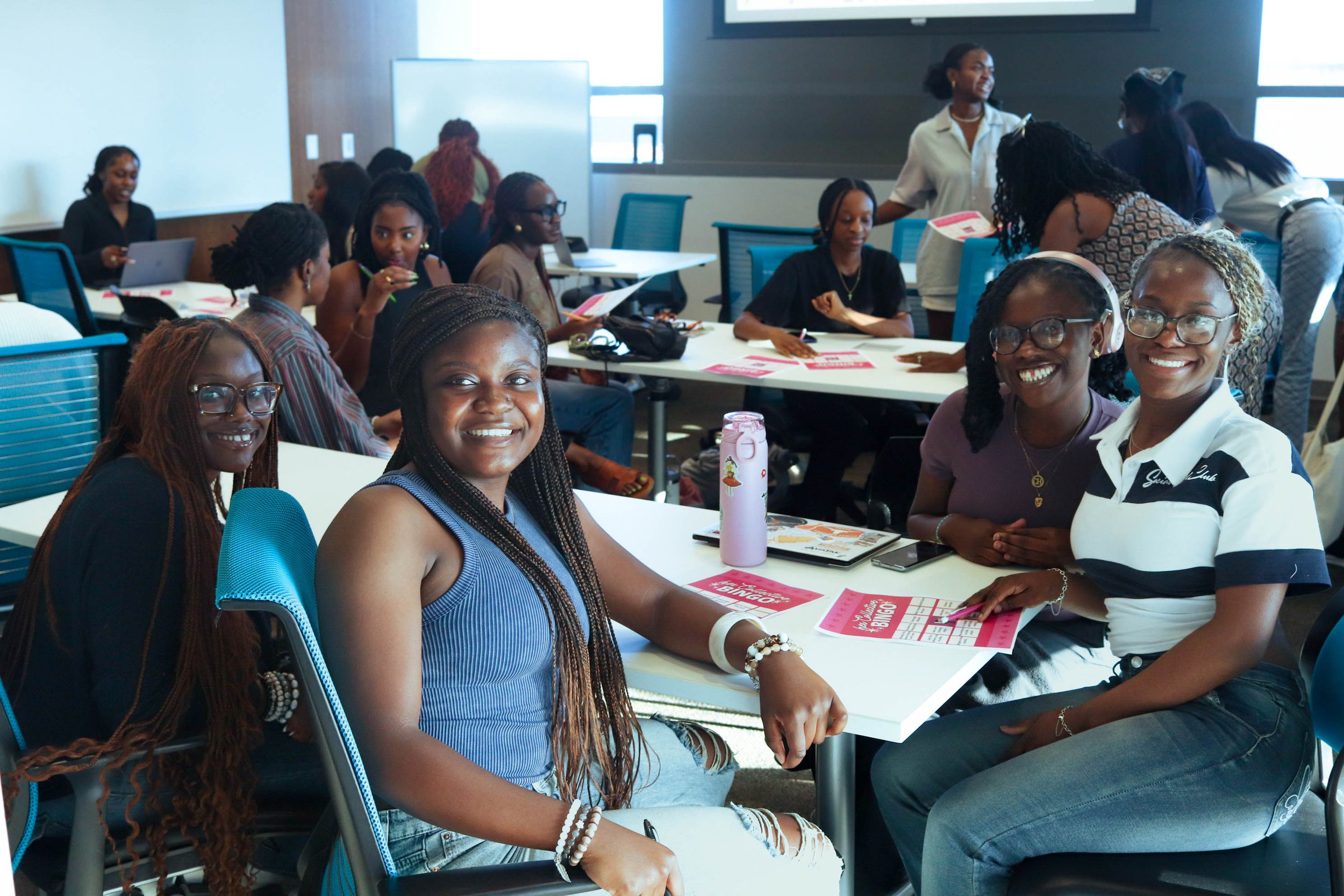 Group of smiling people in a classroom setting sitting around tables with sheets of paper and personal items.