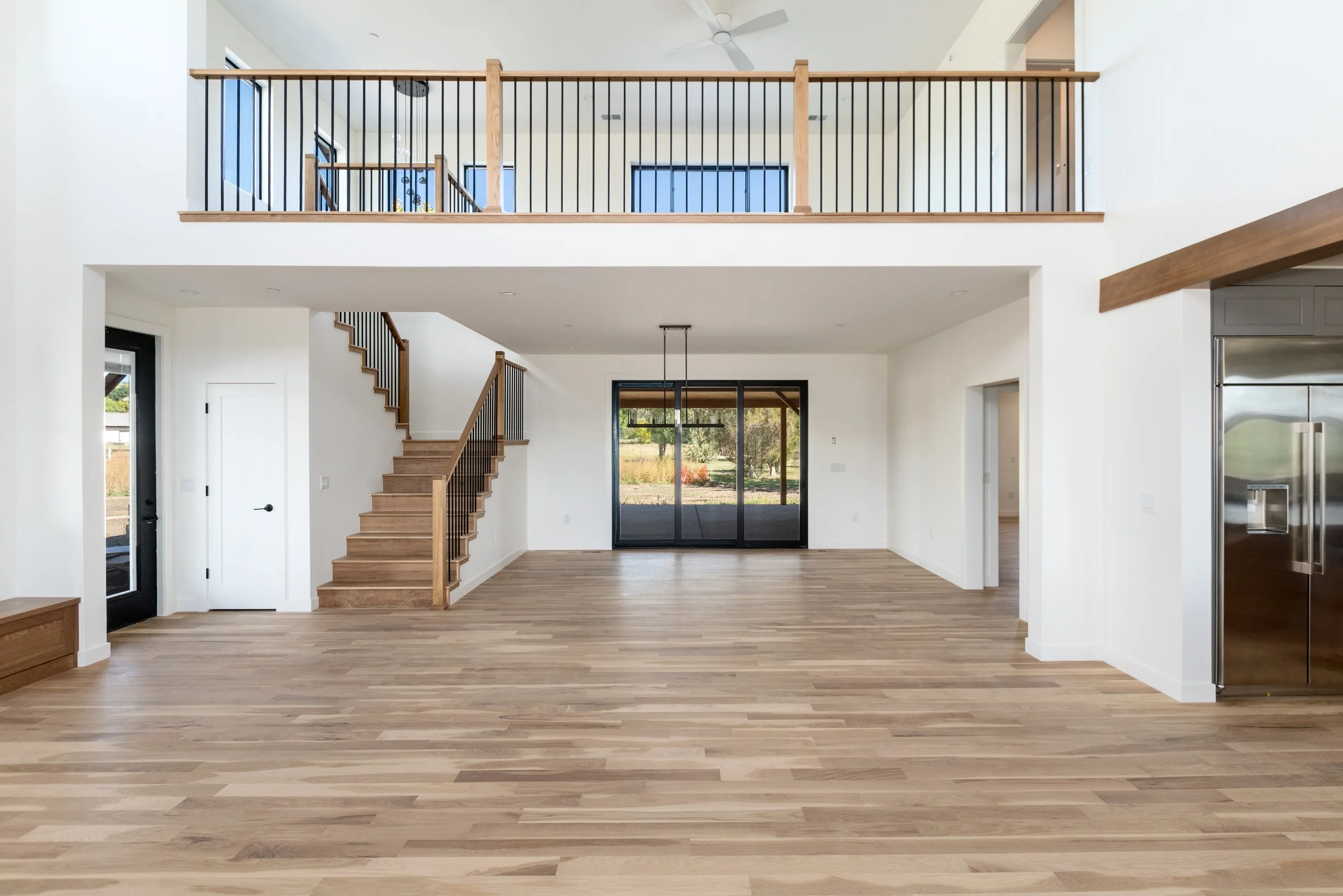 Empty living room with wooden floors, a staircase, and large sliding glass doors leading to an outdoor area with trees.