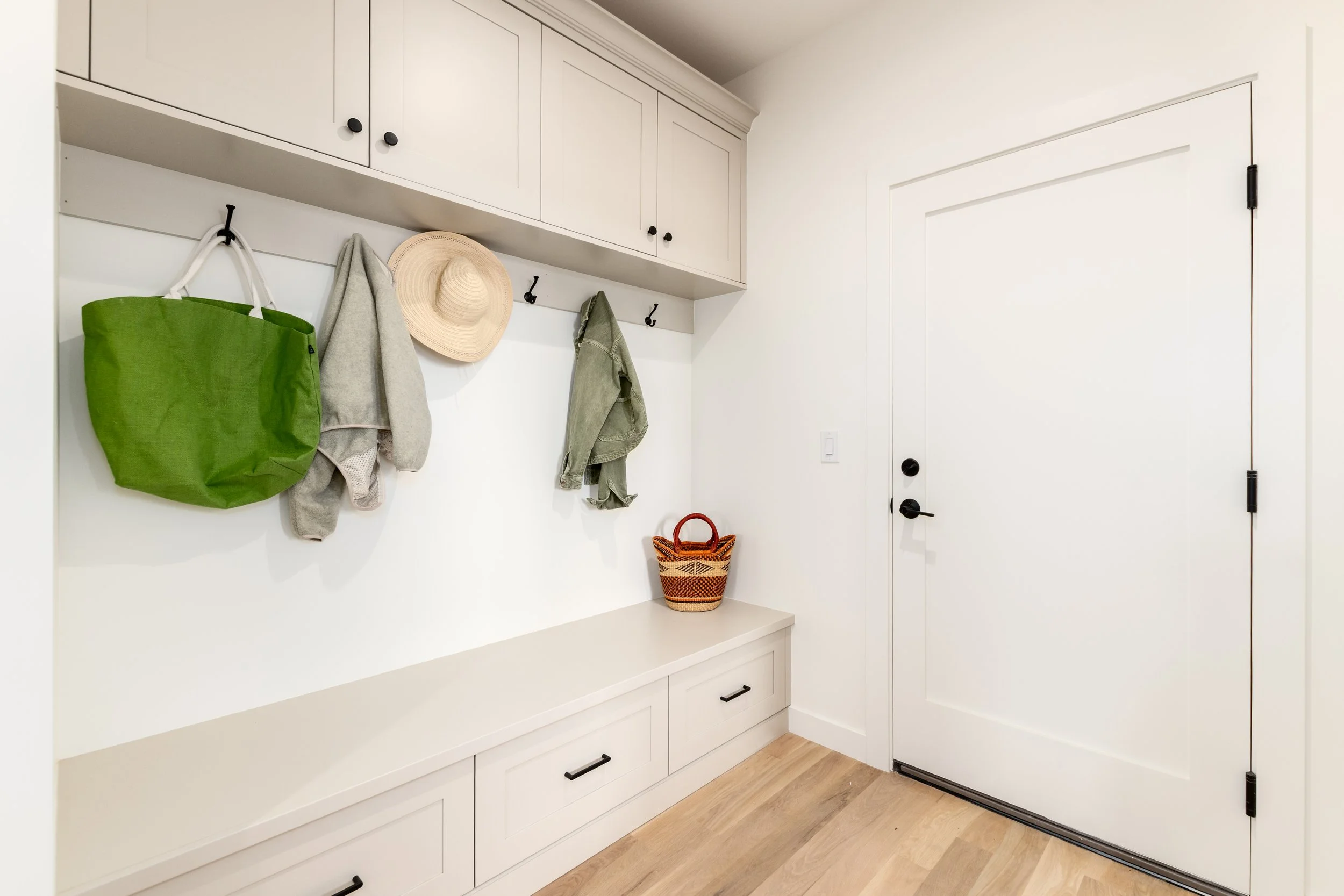 Entryway with white built-in bench and cabinets, black hooks holding a green tote bag, gray hoodie, straw hat, and olive jacket, a woven basket on the bench, and a white door with black hardware.