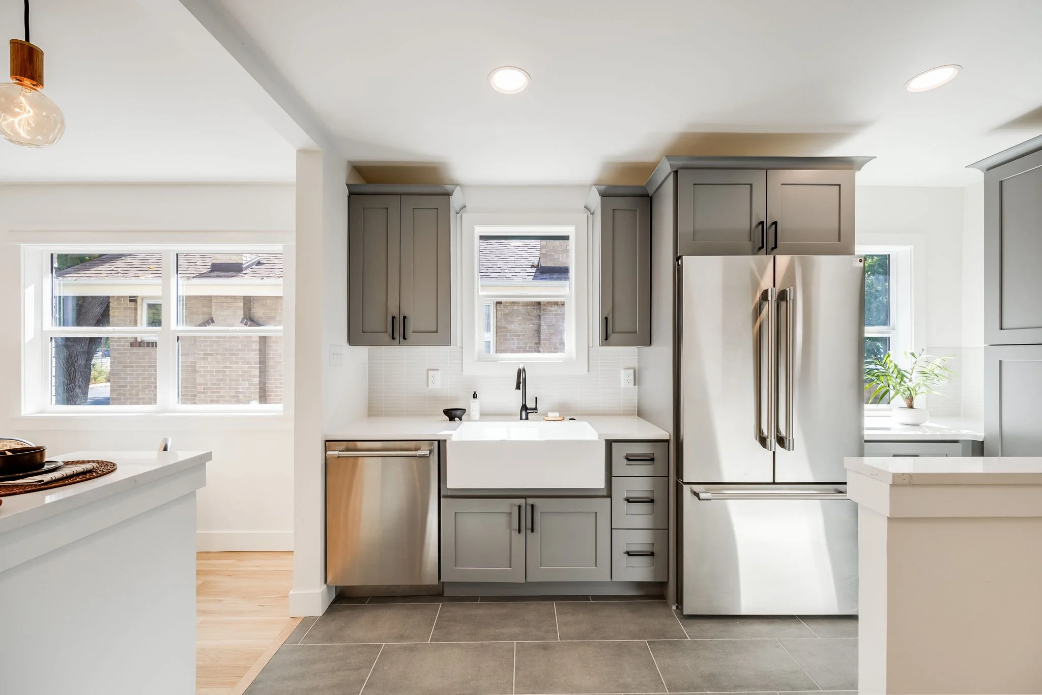 Modern kitchen with gray cabinets, white countertops, stainless steel appliances, a farmhouse sink, and large windows allowing natural light.