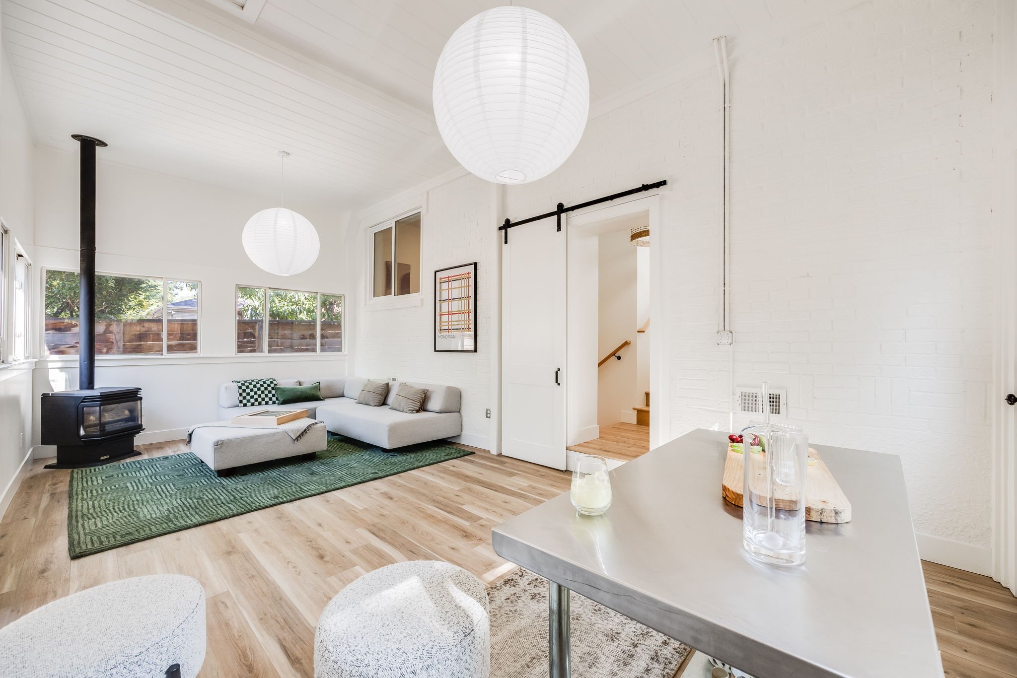 Bright living room with white walls, wooden floors, large windows, a white sectional sofa with green and beige pillows, a green rug, a black wood stove, and a sliding barn door.