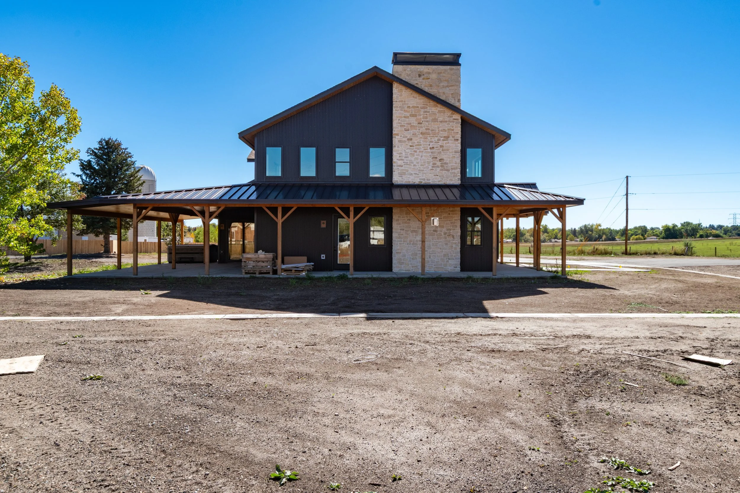 New two-story house under construction with black siding, stone accents, metal roof, and a large covered porch, set in a rural area with open fields and dirt parking lot.