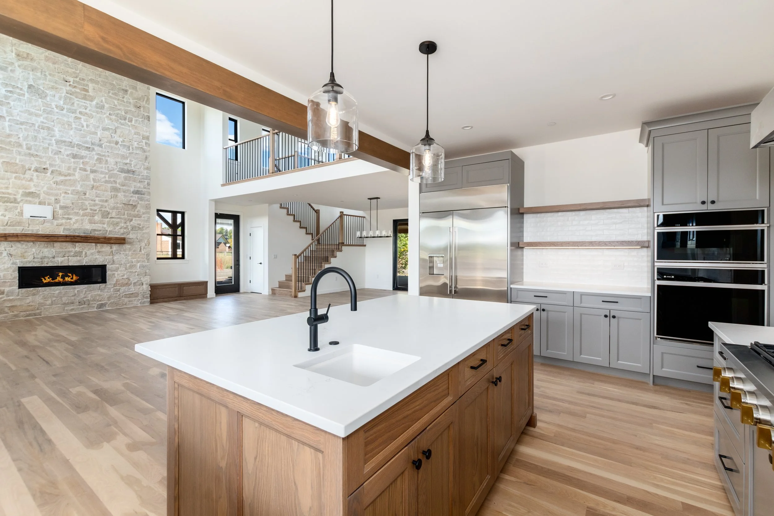 Modern open-concept kitchen with an island, gray cabinetry, stainless steel refrigerator, and pendant lighting, with a living area and staircase in the background.