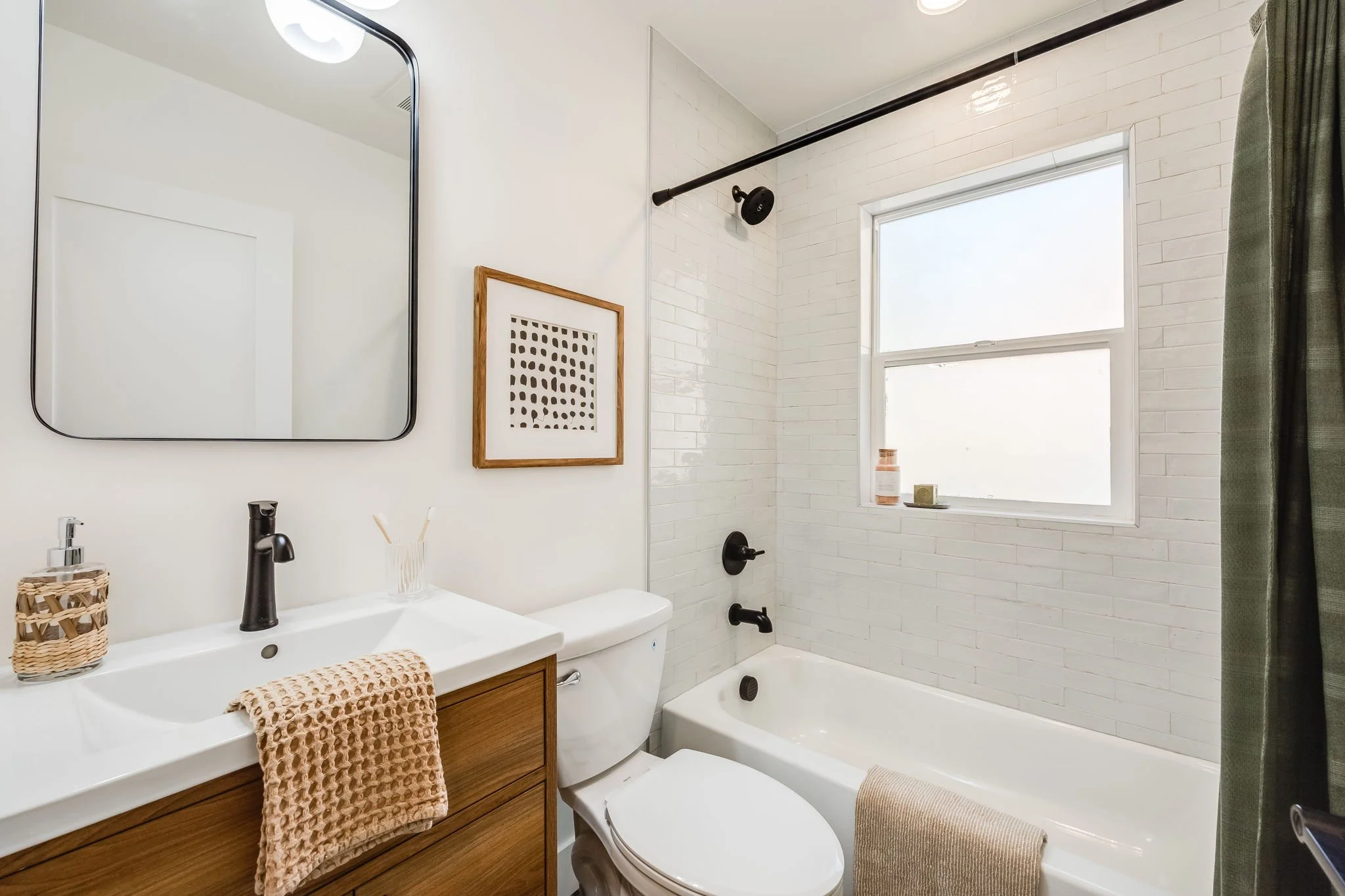 A modern bathroom with a white bathtub, black fixtures, white brick wall tiles, a window with frosted glass, a wooden vanity with a white sink, a mirror, a framed abstract art piece, and a green shower curtain.
