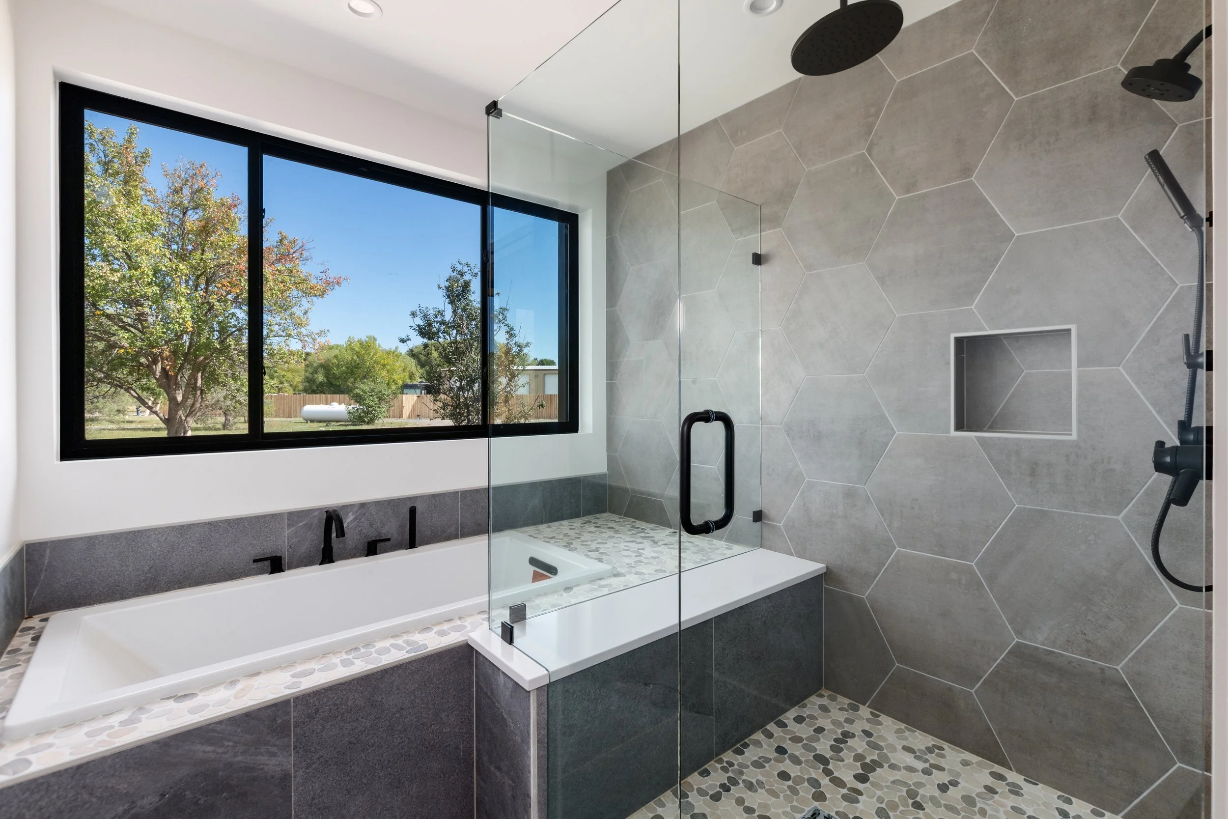 Modern bathroom with large window, white bathtub, and walk-in shower with hexagonal tiles, black fixtures, and pebble floor.