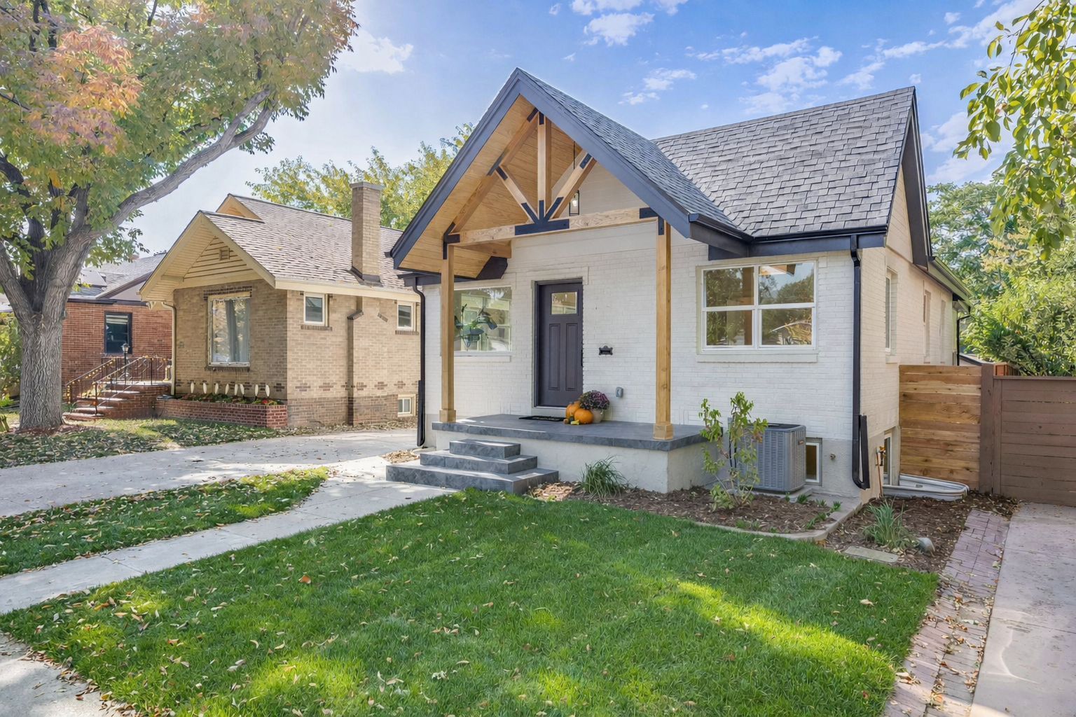 A white brick house with a black door and a small front porch, decorated with pumpkins and flowers, has a gabled roof and is surrounded by a green lawn and trees.