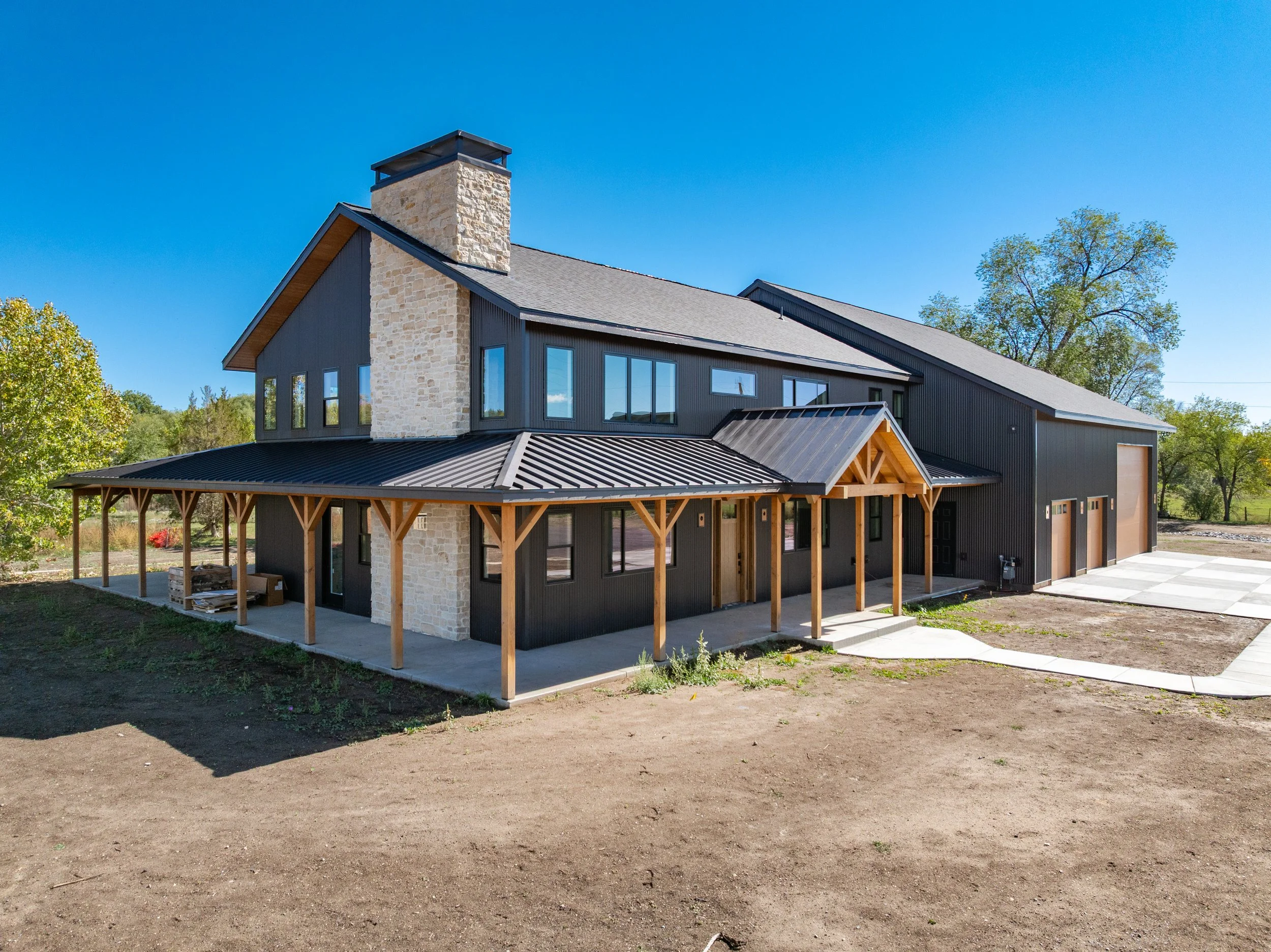 Modern two-story house with black exterior siding, a stone chimney, and a metal roof with a porch, set in a rural area with trees and clear blue sky.