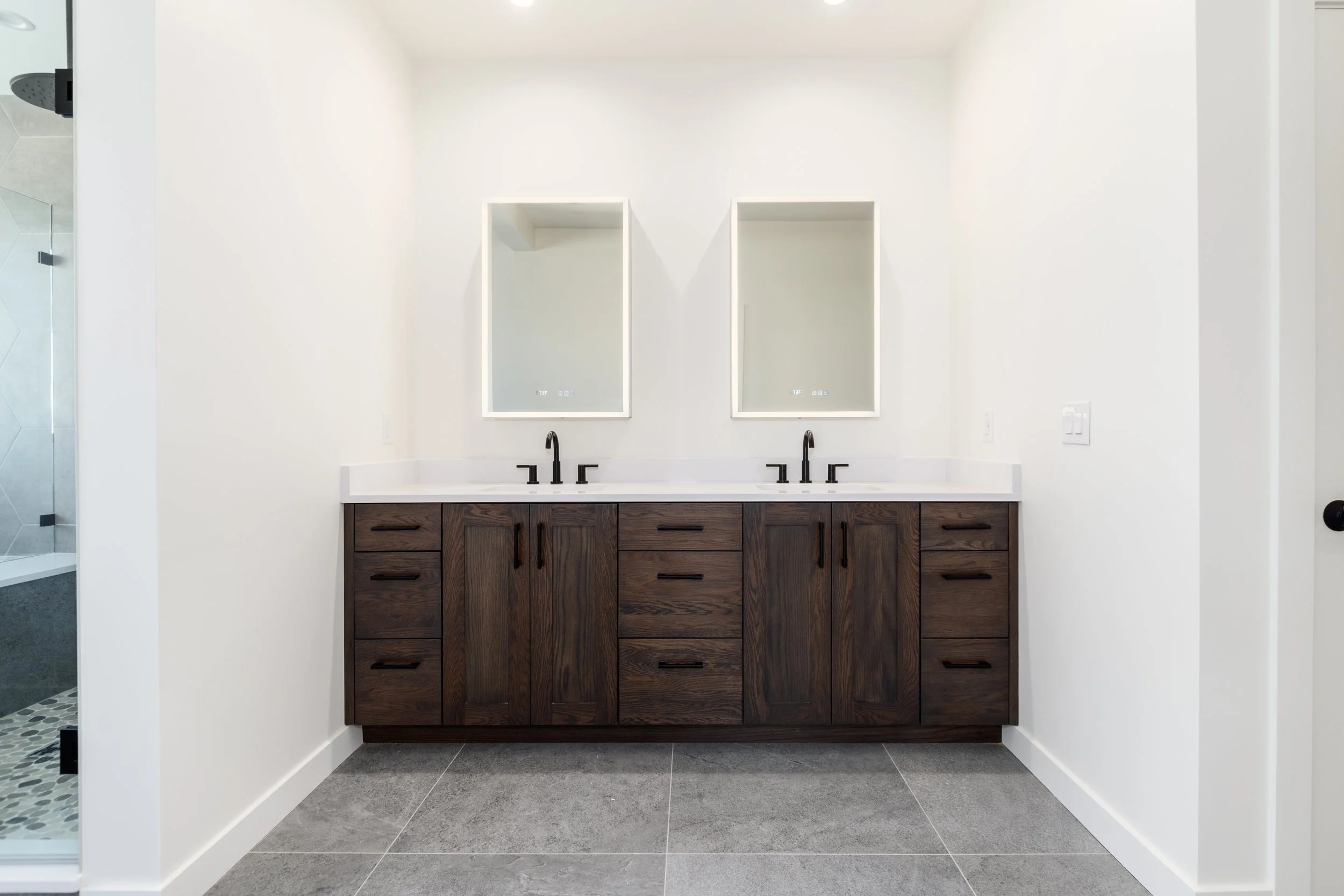 Double vanity bathroom with dark wood cabinets, white countertop, two black faucets, two illuminated rectangular mirrors, and a walk-in shower visible on the left with glass door and tiled walls.