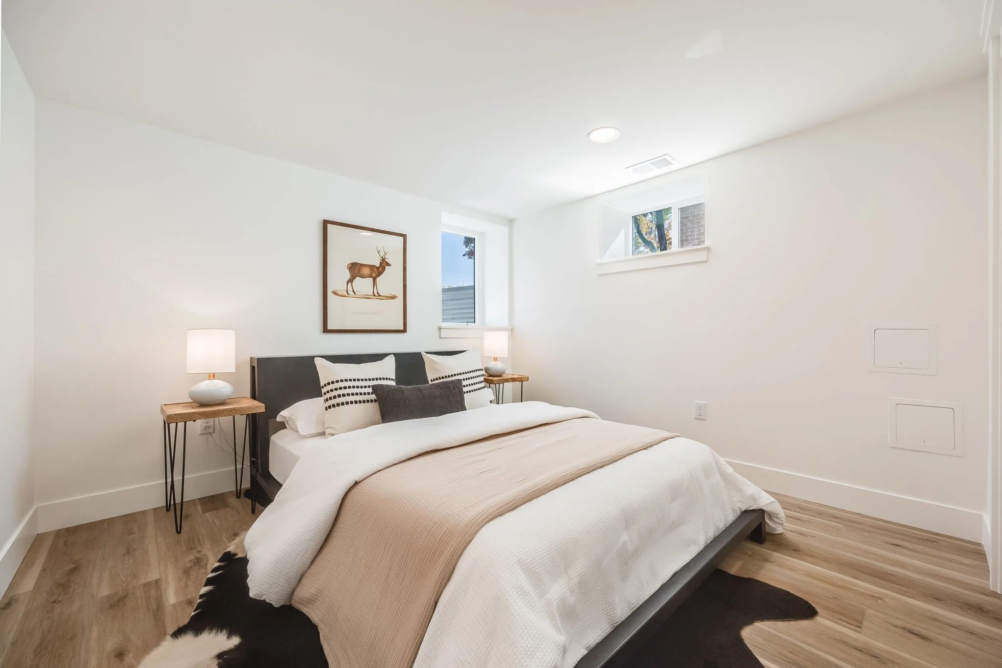 A minimalistic bedroom with a bed, two nightstands with lamps, a framed picture of a deer on the wall, and small windows letting in natural light, with hardwood flooring.