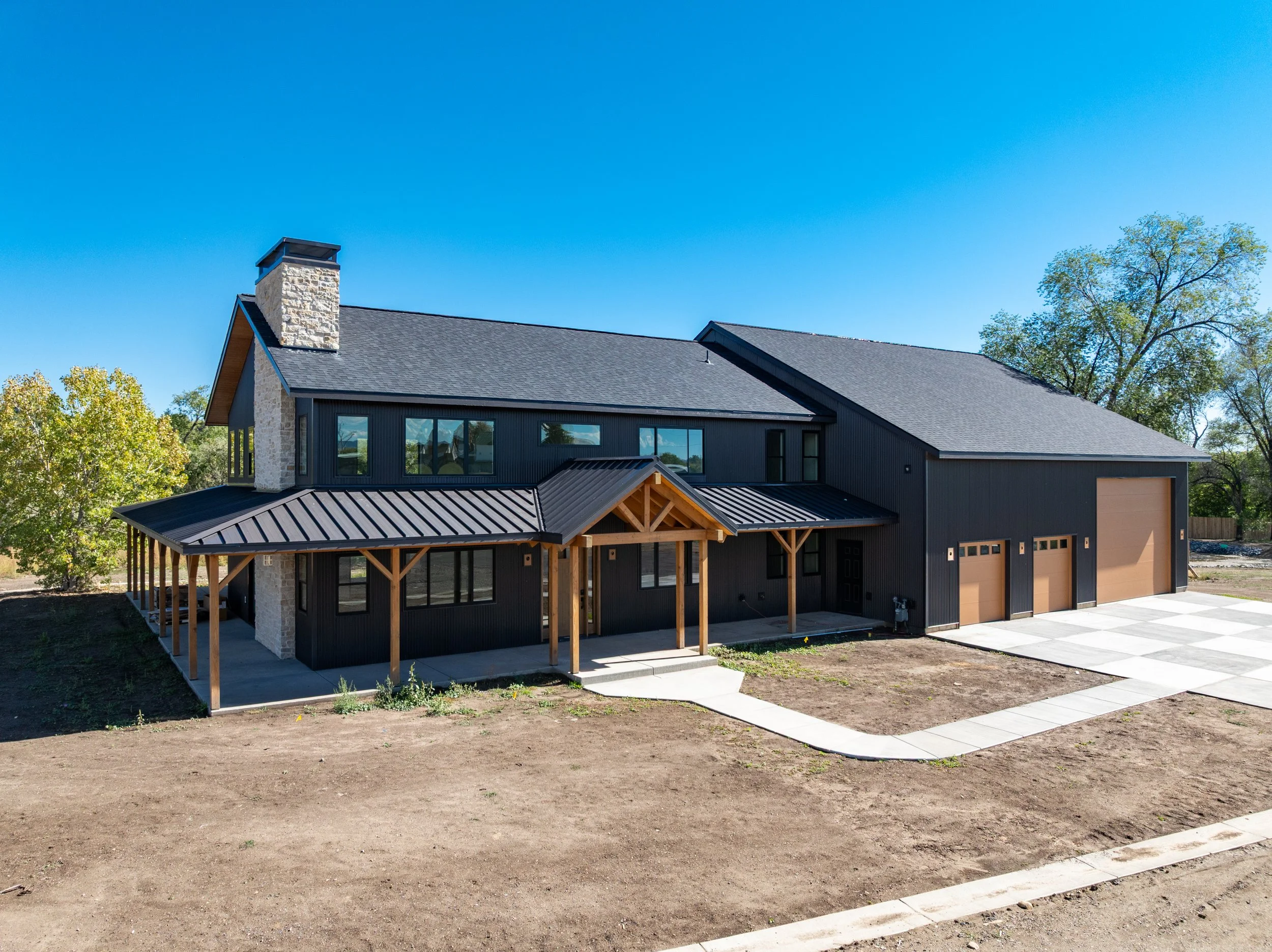 Modern two-story house with black exterior, large windows, a covered porch with wooden beams, a chimney, and three garage doors on a clear day.