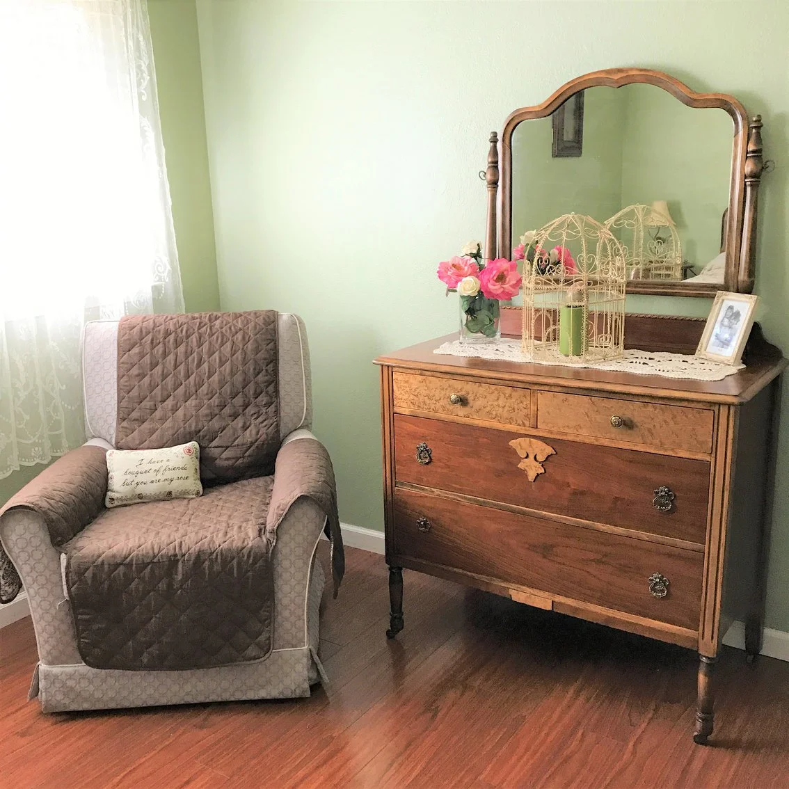 A vintage dresser with a mirror, decorated with a vase of pink and white flowers, a framed photograph, and a birdcage, next to an upholstered armchair with a quilted cover and a small decorative pillow, in a room with green walls and hardwood floors.