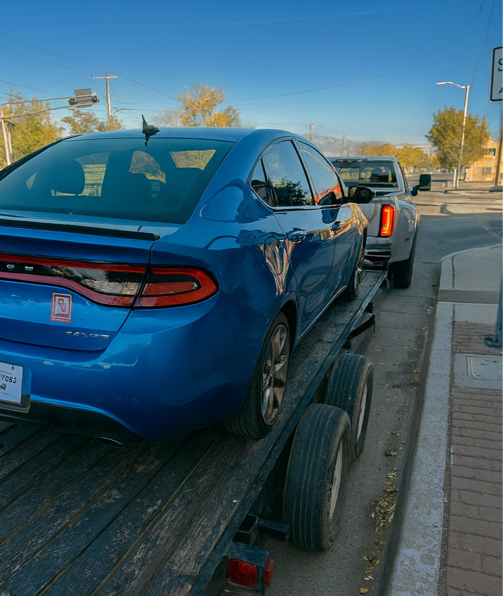 A blue sedan being transported on a flatbed truck, with a pickup truck connected to it in the background. The scene is on a city street with trees, utility poles, and a traffic signal visible.