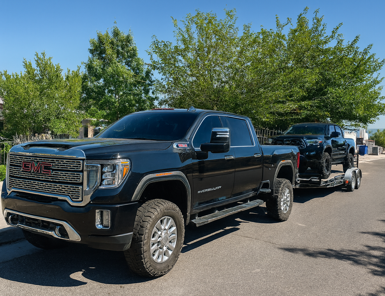 Black GMC pickup truck towing a trailer with a black vehicle and equipment on a suburban street with green trees and blue sky.