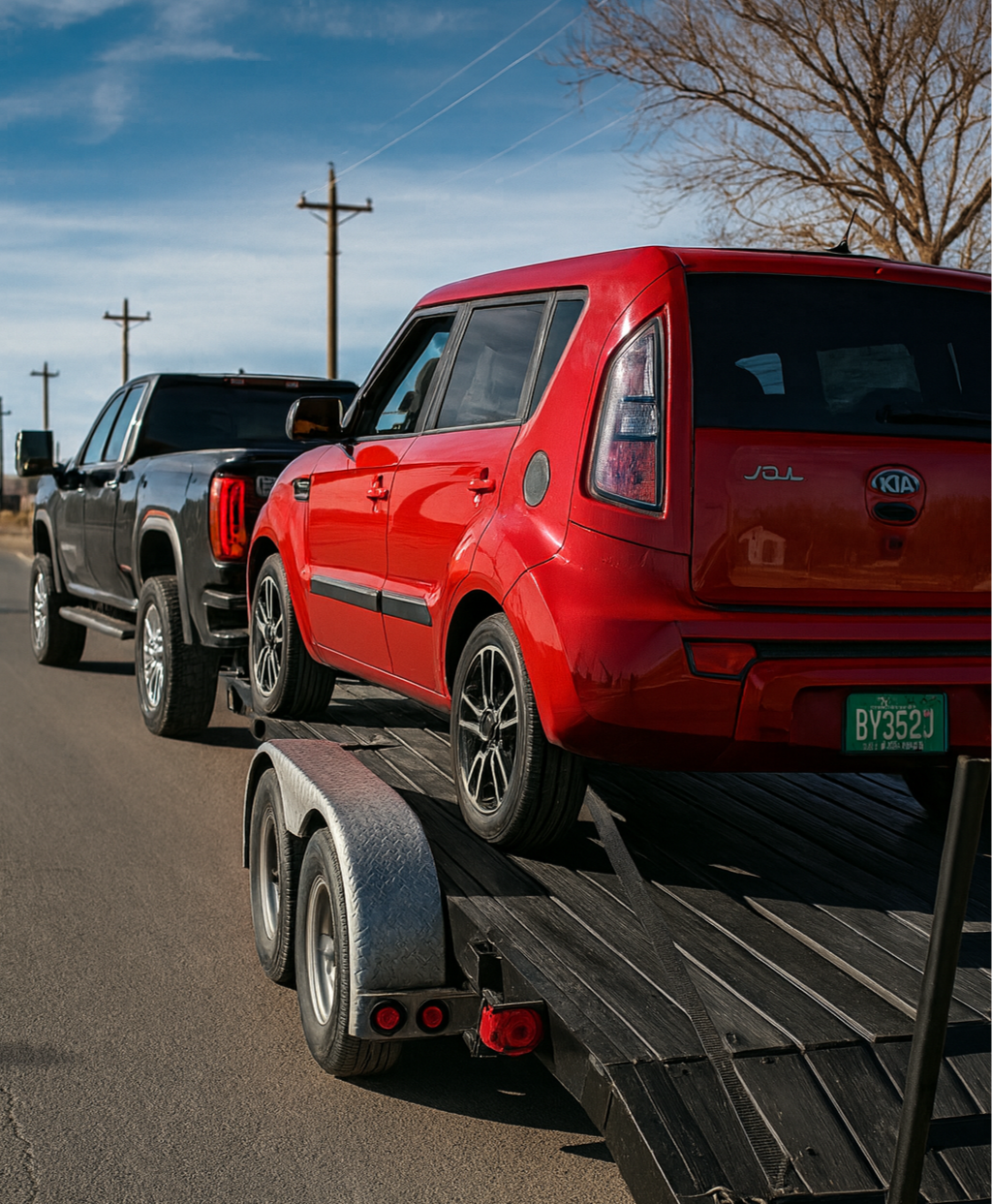 A red Kia Soul being transported on a flatbed trailer attached to a black pickup truck.