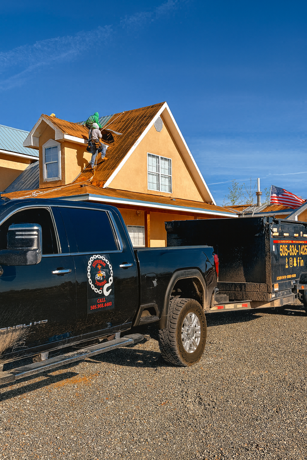 Workers are installing a new roof on a yellow house using a black pickup truck with a trailer, in a residential area under a clear blue sky, with an American flag visible.