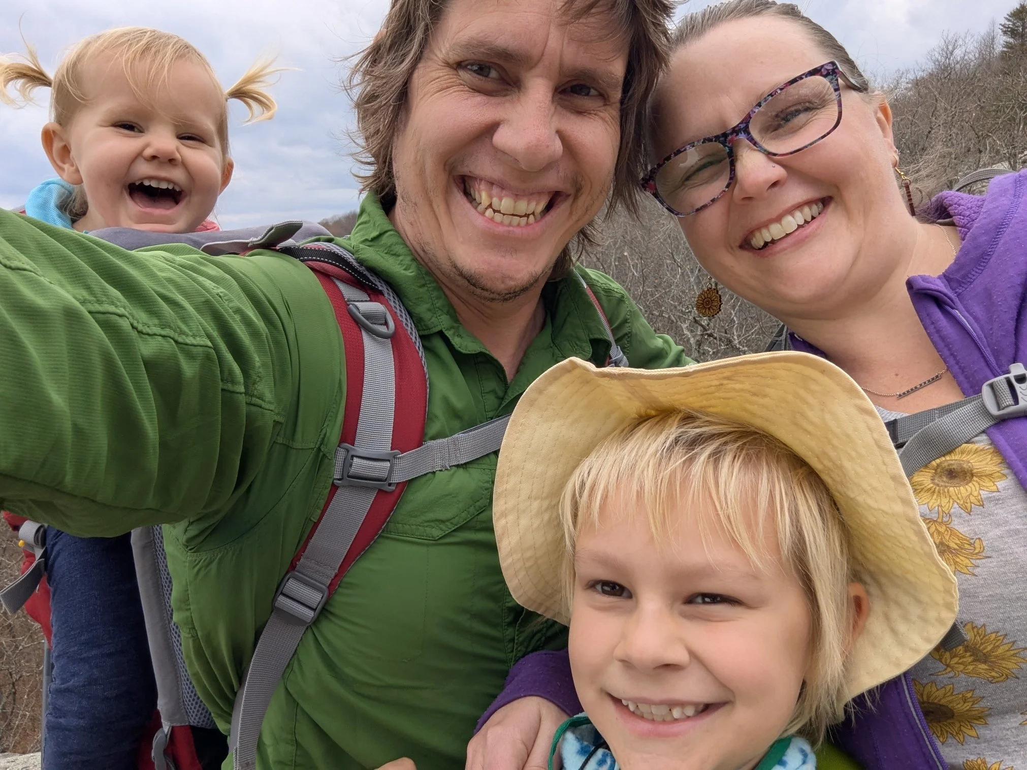 A smiling family of four taking a selfie outdoors, with a father, a mother wearing glasses, a young girl with a hat, and a young boy with blonde hair, all appearing happy and enjoying nature.