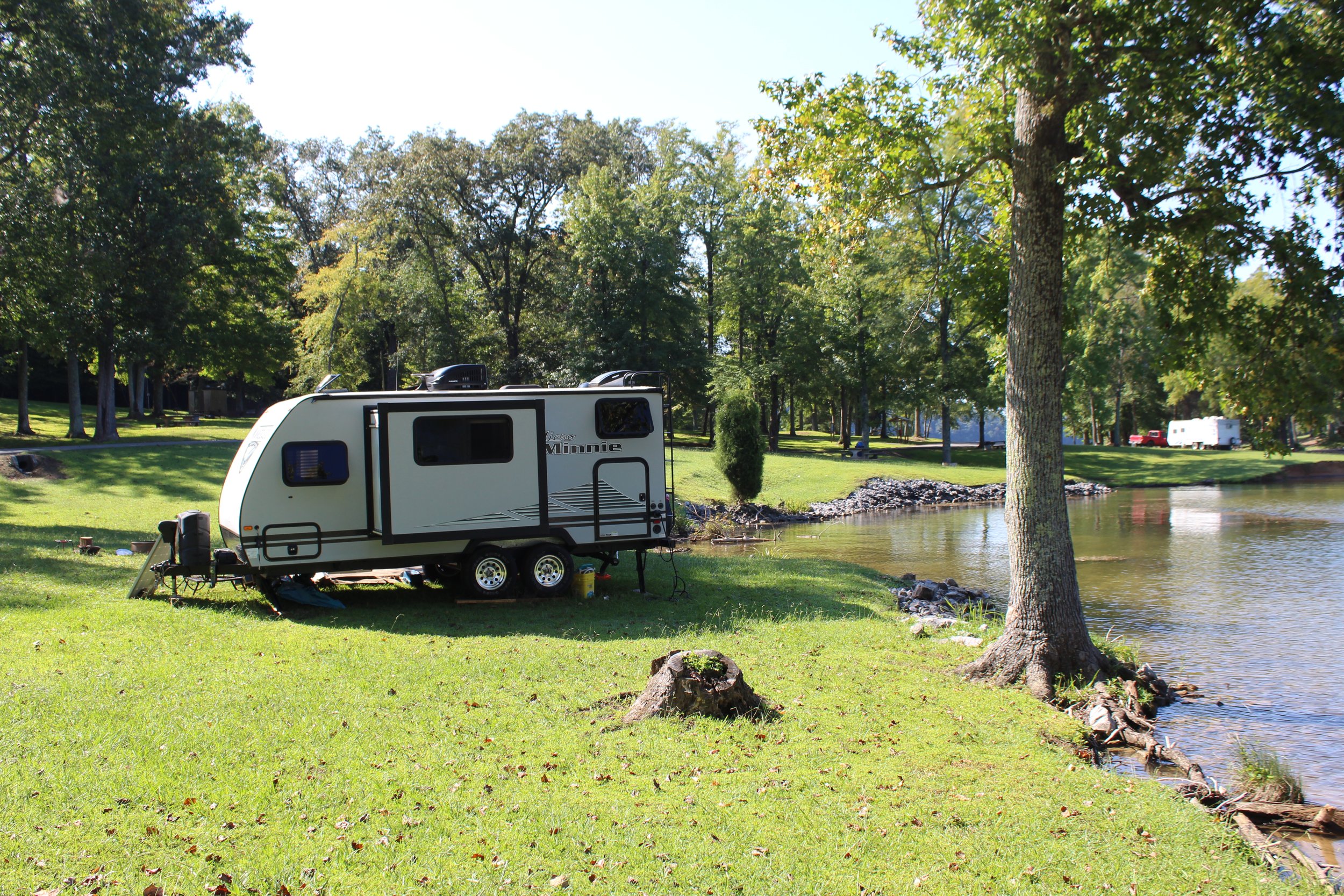 A white travel trailer parked on a grassy area near a lake with trees in the background.
