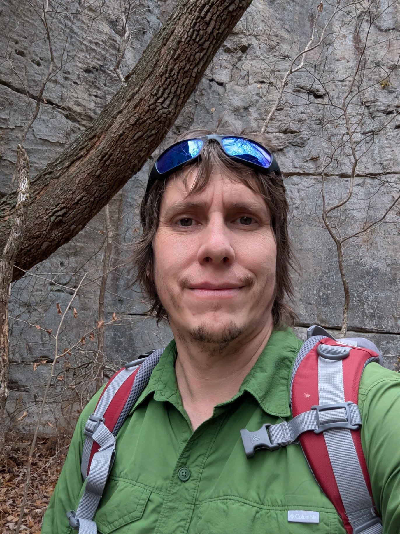A man taking a selfie outdoors in a wooded area, wearing a green shirt, sunglasses on his head, and a red backpack. Behind him is a rock face and trees with bare branches.