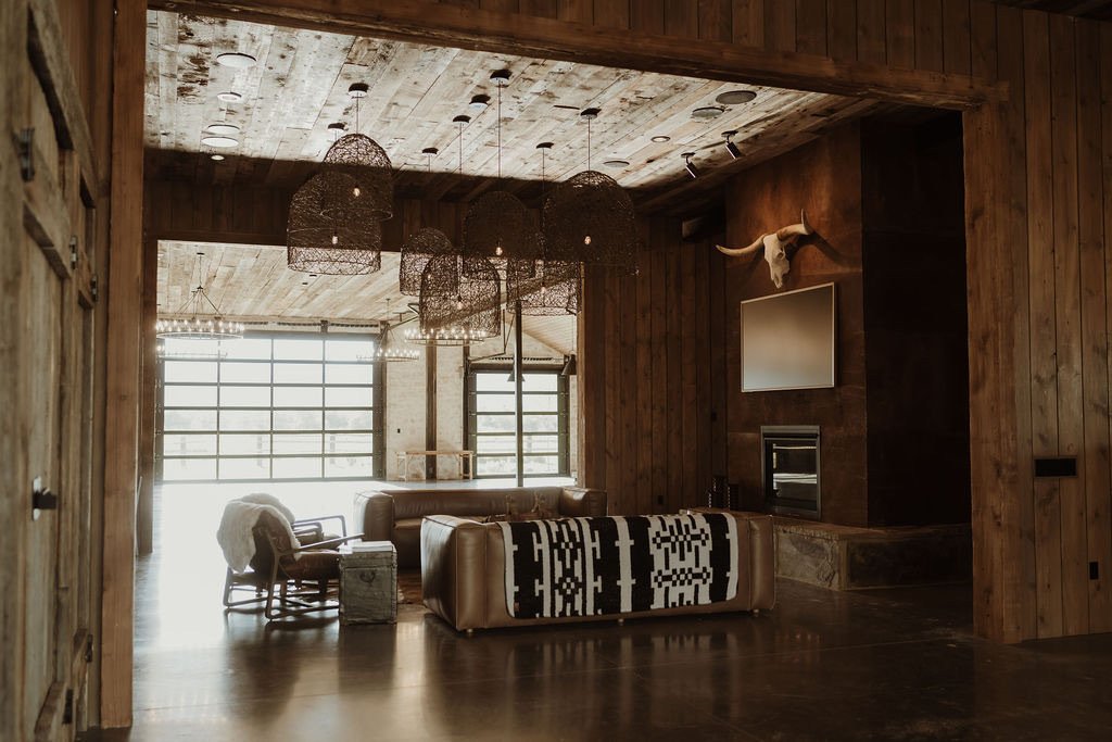 A rustic living room with wood-paneled walls and ceiling, featuring large windows, a leather sofa with a black and white patterned blanket, wooden chairs with cushions, a stone fireplace, and decorative animal skulls on the wall.