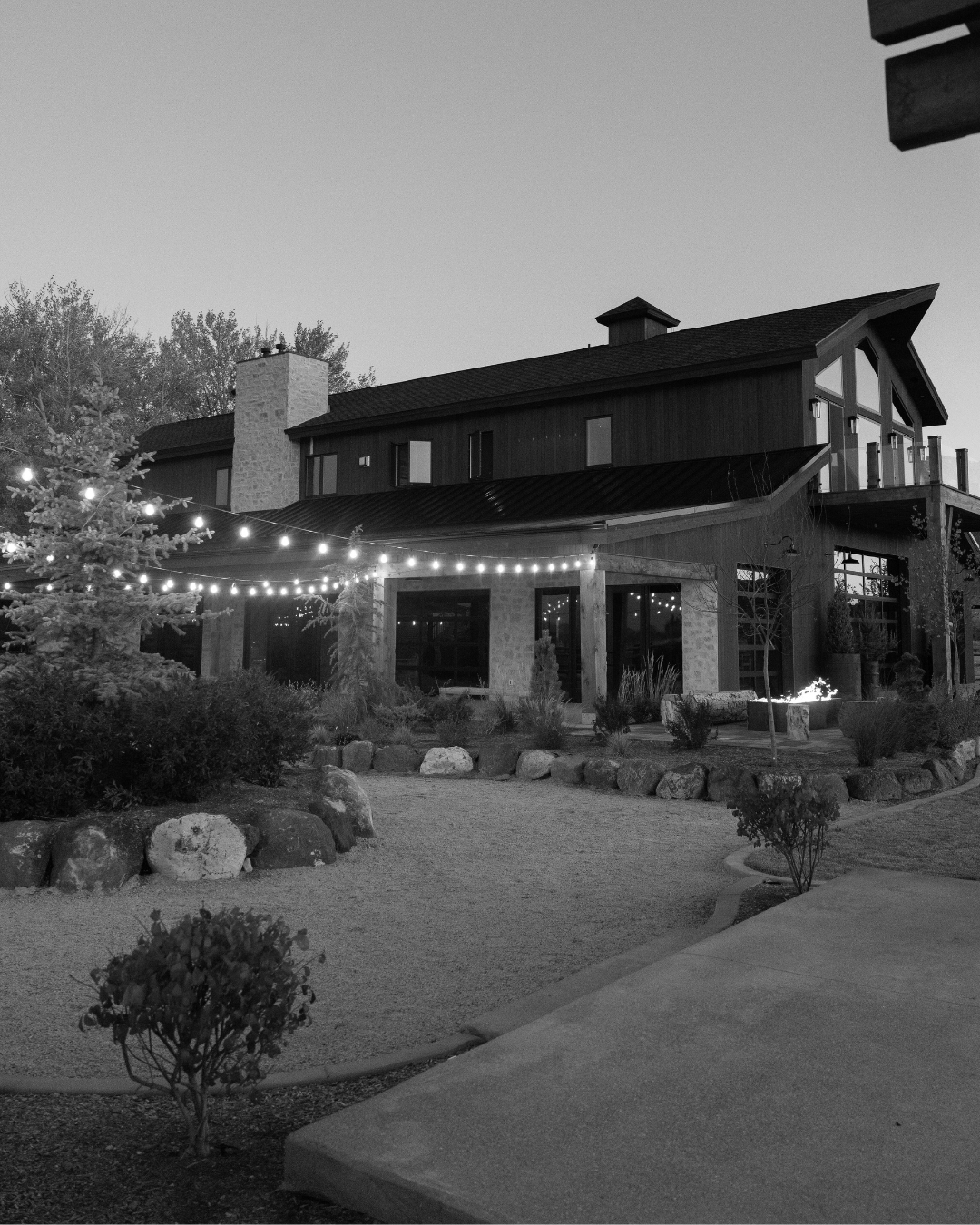 A black and white photo of a modern two-story house with large windows and a chimney, decorated with string lights in the yard, surrounded by plants and trees.