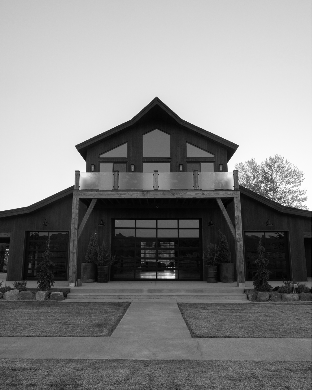 Black and white photo of a modern barn-style building with large windows and a balcony, surrounded by a lawn and trees.