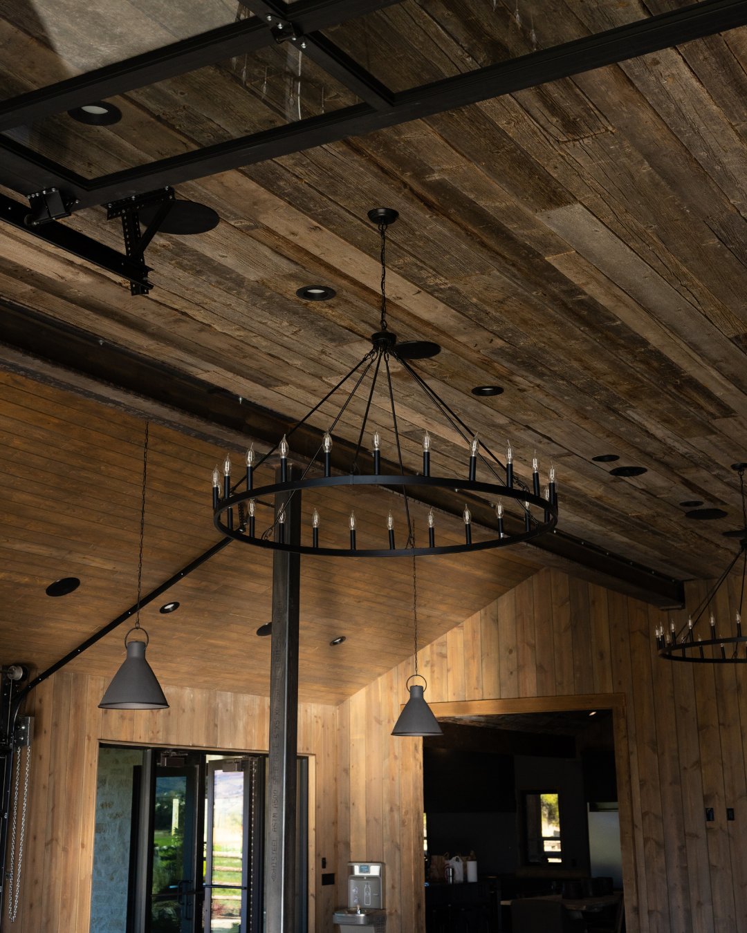Interior of a room with a wooden ceiling, black circular chandelier with candle-style lights, hanging grey pendant lights, and wooden walls.