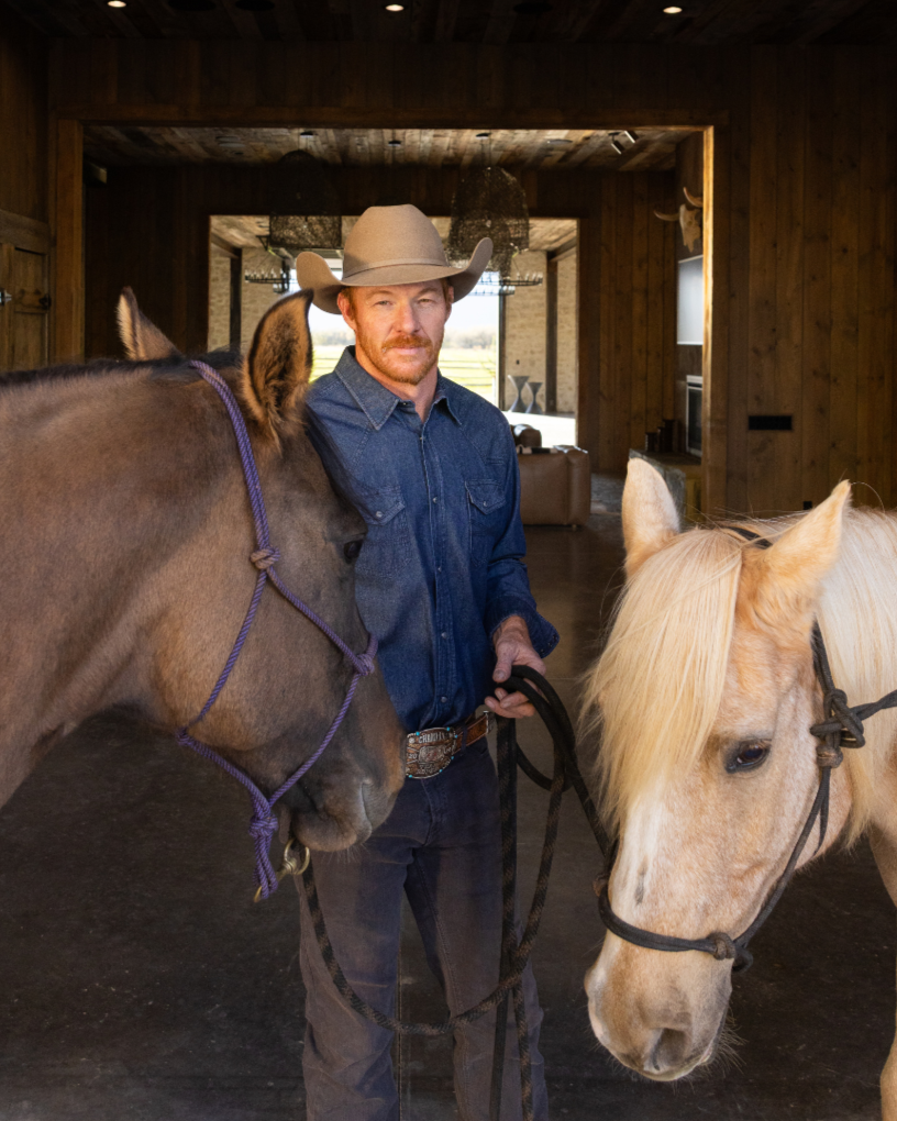 A man wearing a cowboy hat and a denim shirt standing indoors with two horses, one brown and one light blonde, in a wooden room with large windows and modern lighting fixtures.