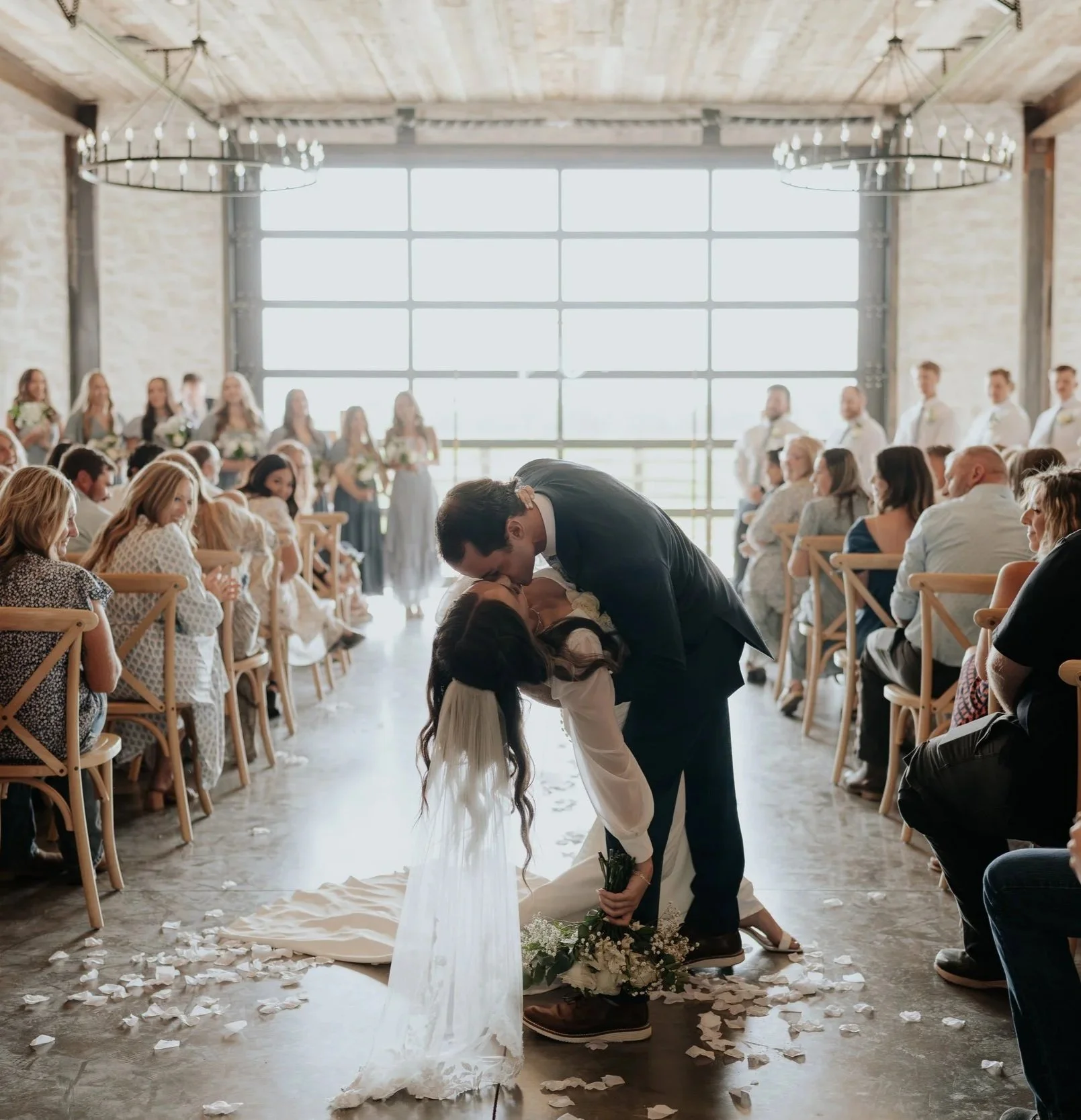 A man in a suit and a woman in a wedding dress are kissing during a wedding ceremony in a bright, rustic indoor venue with large windows, wood ceiling, and large chandeliers. The groom is holding the bride in an embrace as she kneels on the floor, and there are flower petals scattered on the ground.