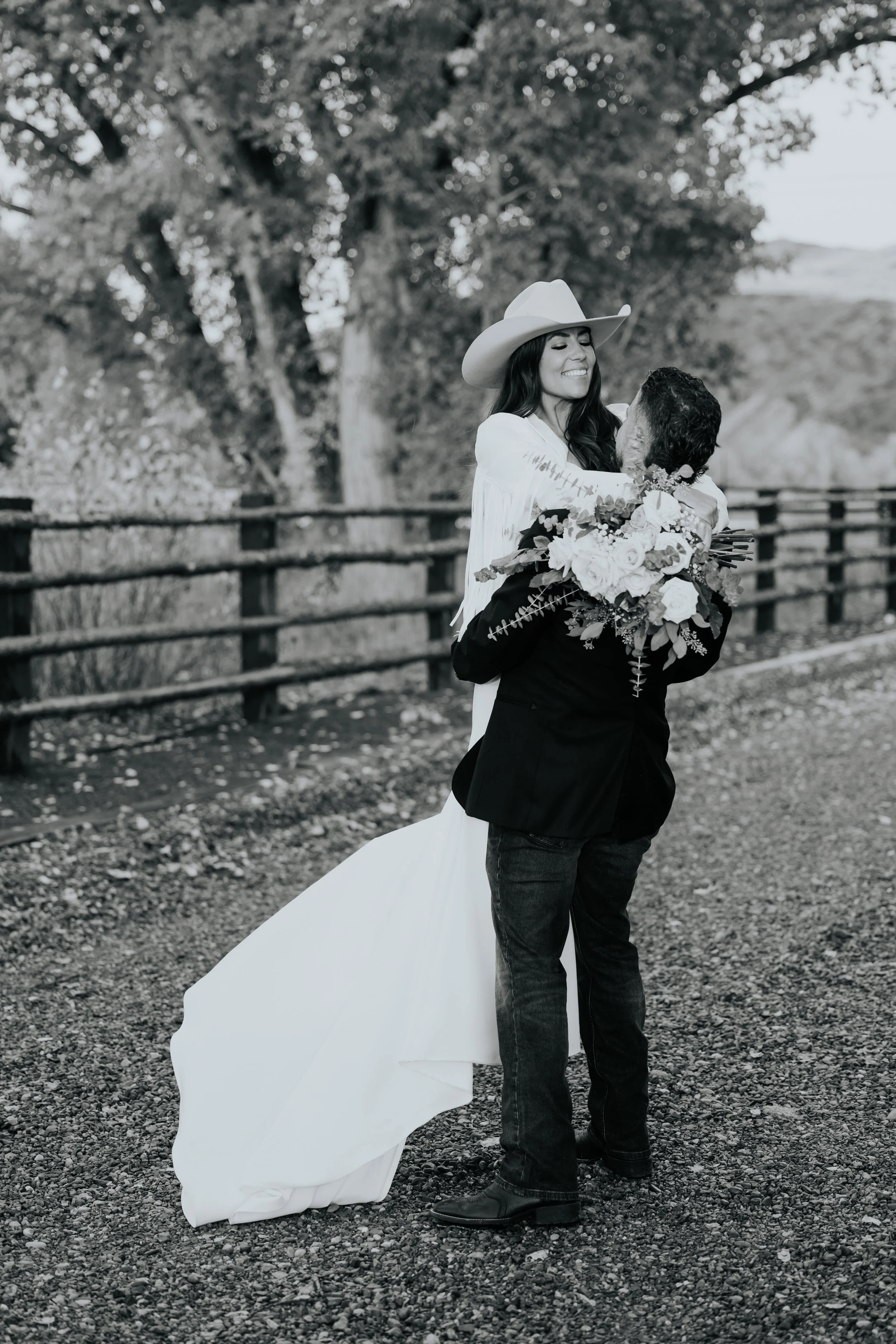 A woman in a white dress and cowboy hat is smiling and embracing a man holding a bouquet of flowers. They are outdoors with trees and a wooden fence in the background.