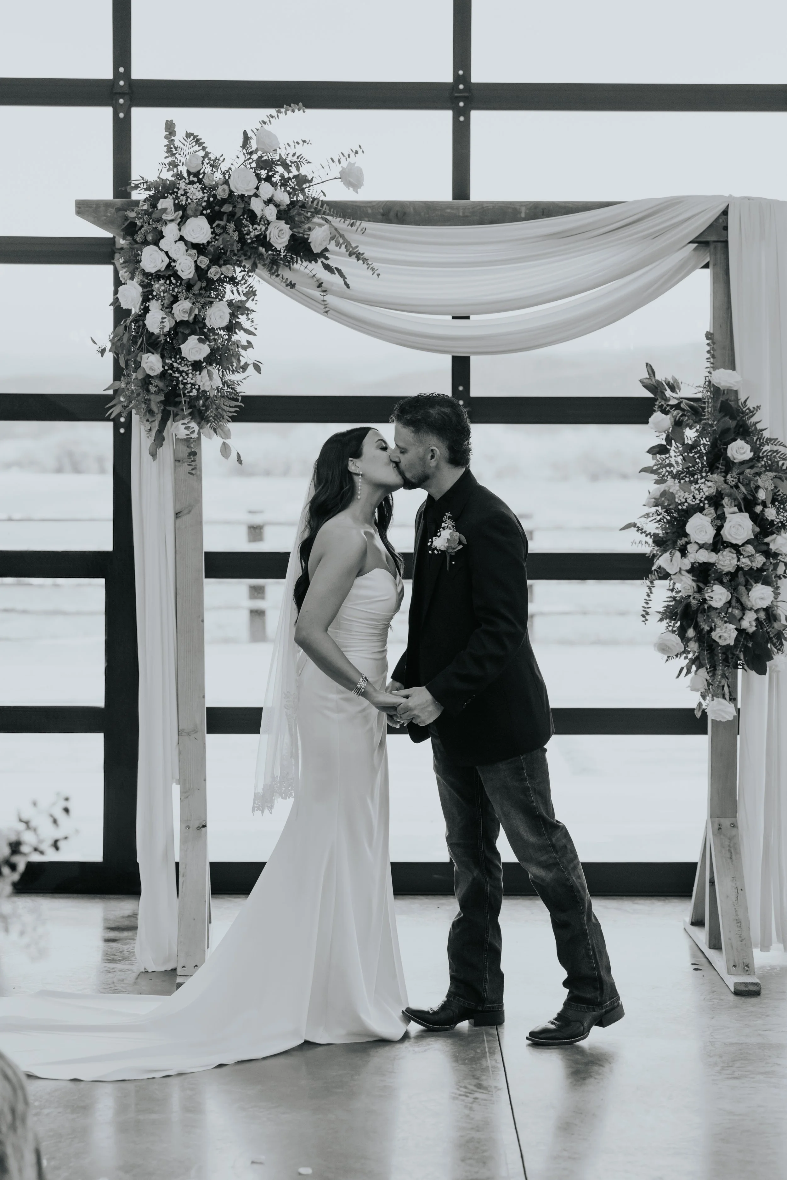 A bride and groom sharing a kiss during their wedding ceremony in front of a floral altar with draped fabric, set against a glass window background.