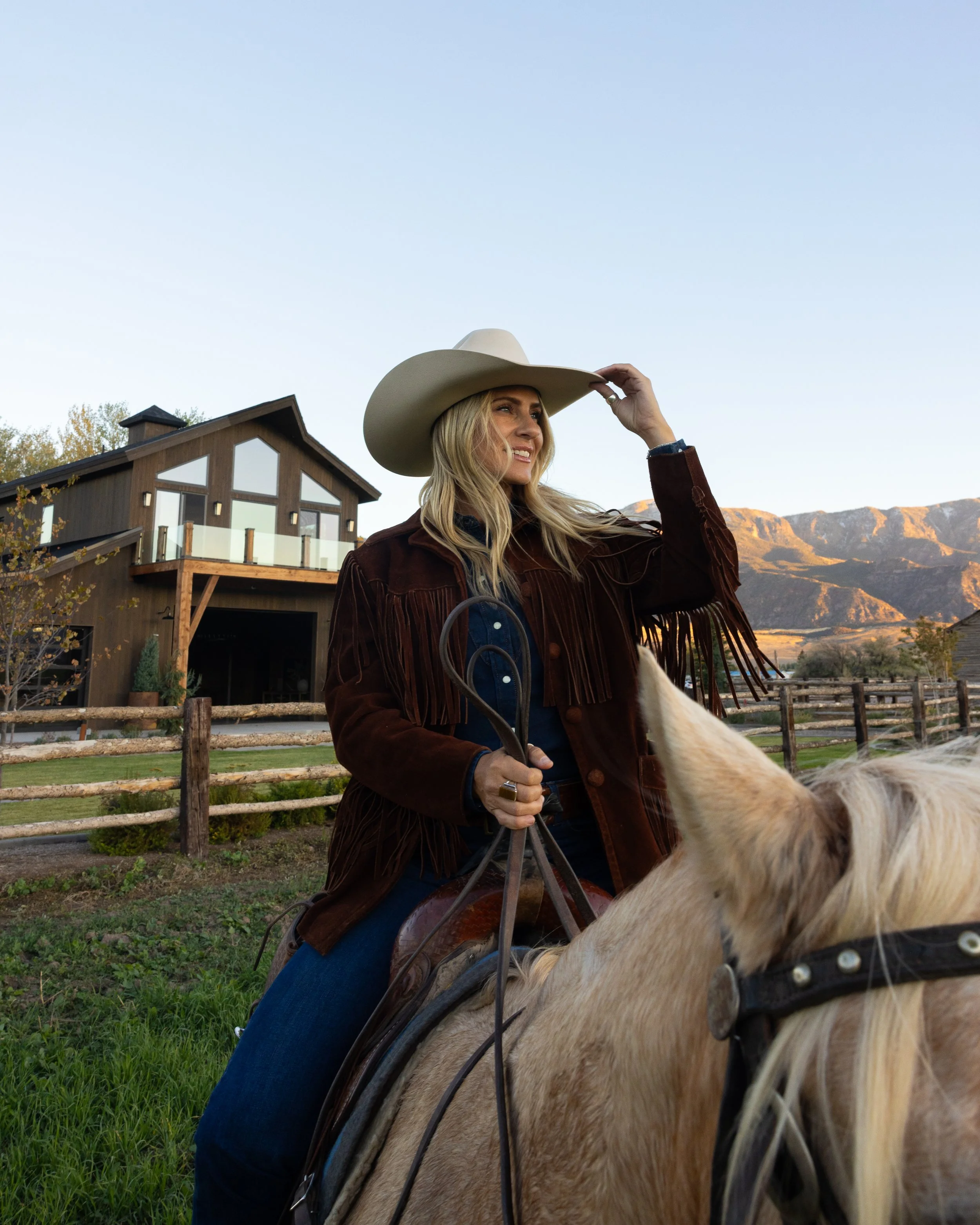 A woman wearing a cowboy hat riding a horse in a rural setting with a house and mountains in the background.