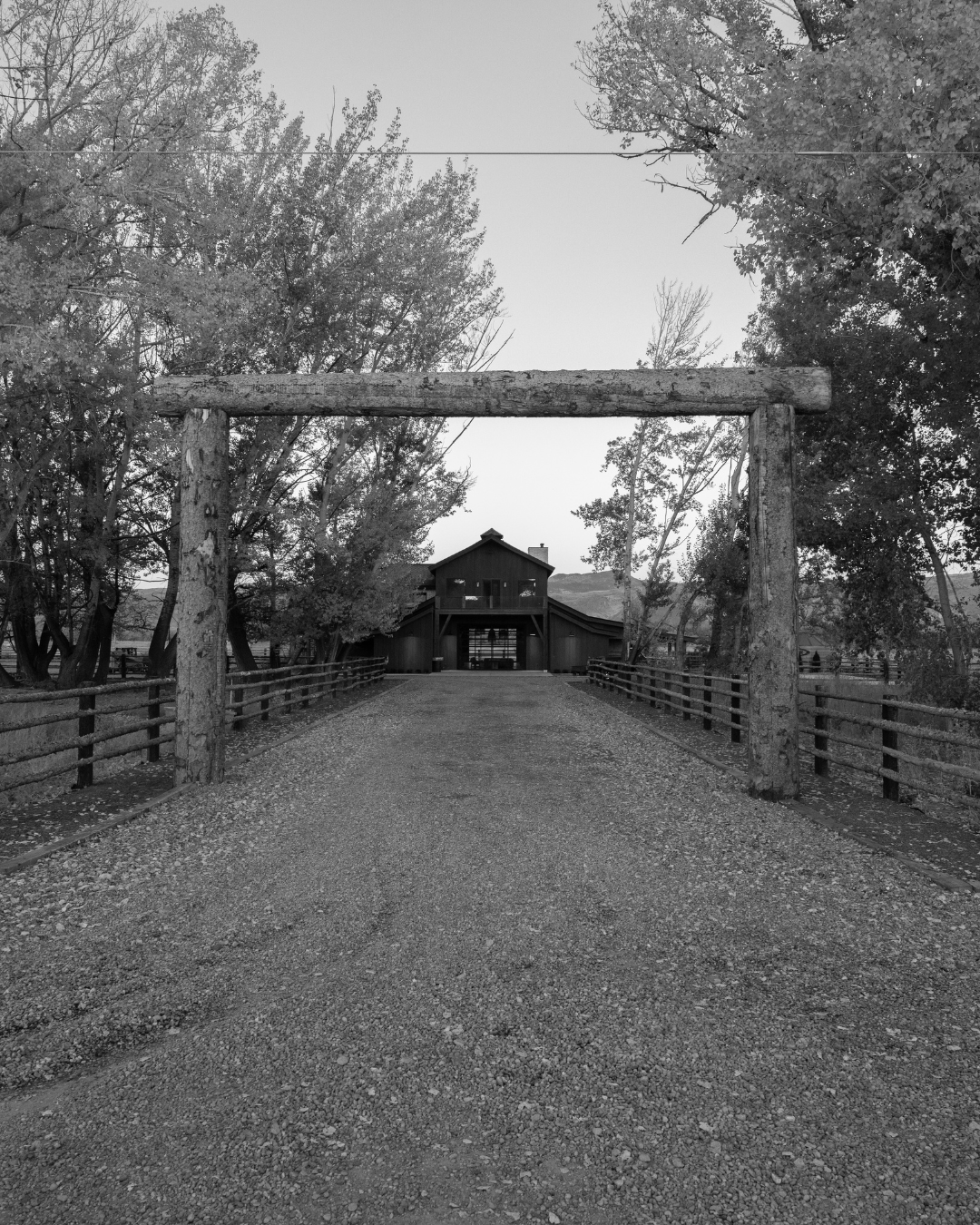 A gravel driveway leading to a large wooden barn, framed by a rustic log gate and fenced with wooden rails, surrounded by trees and open fields.