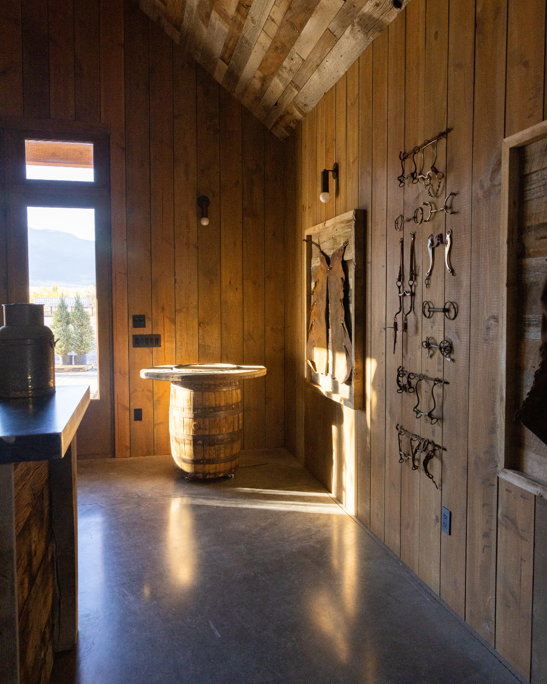 Wood-paneled interior of a rustic room with a wooden barrel table and various old tools and decorations on the wall, sunlight streaming through a window.