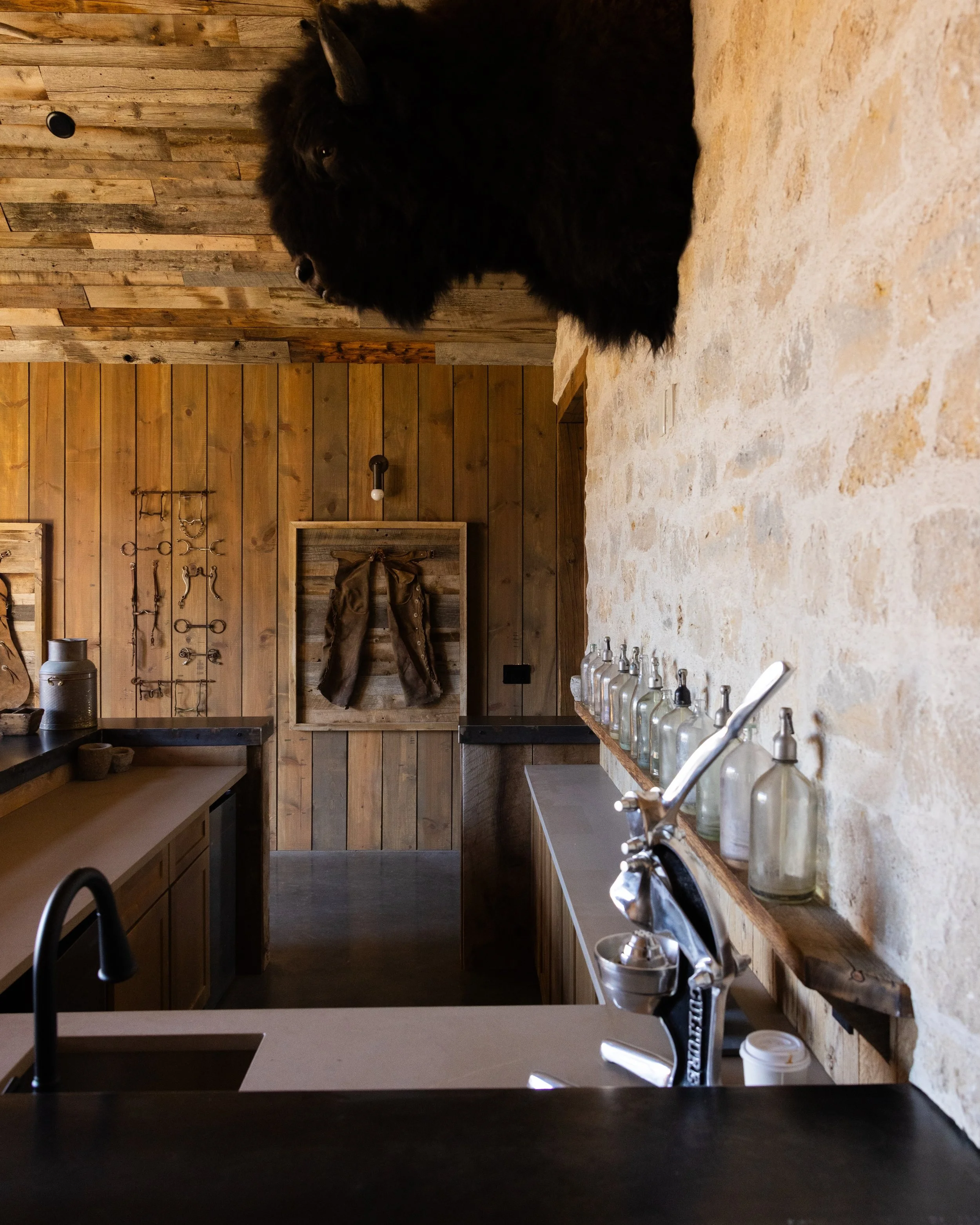 A rustic kitchen with wooden walls and a white stone countertop. Several glass bottles with pumps are lined up on the counter. There is a black sink and faucet visible, along with kitchen utensils hanging on hooks and a cabinet with a pair of work gl