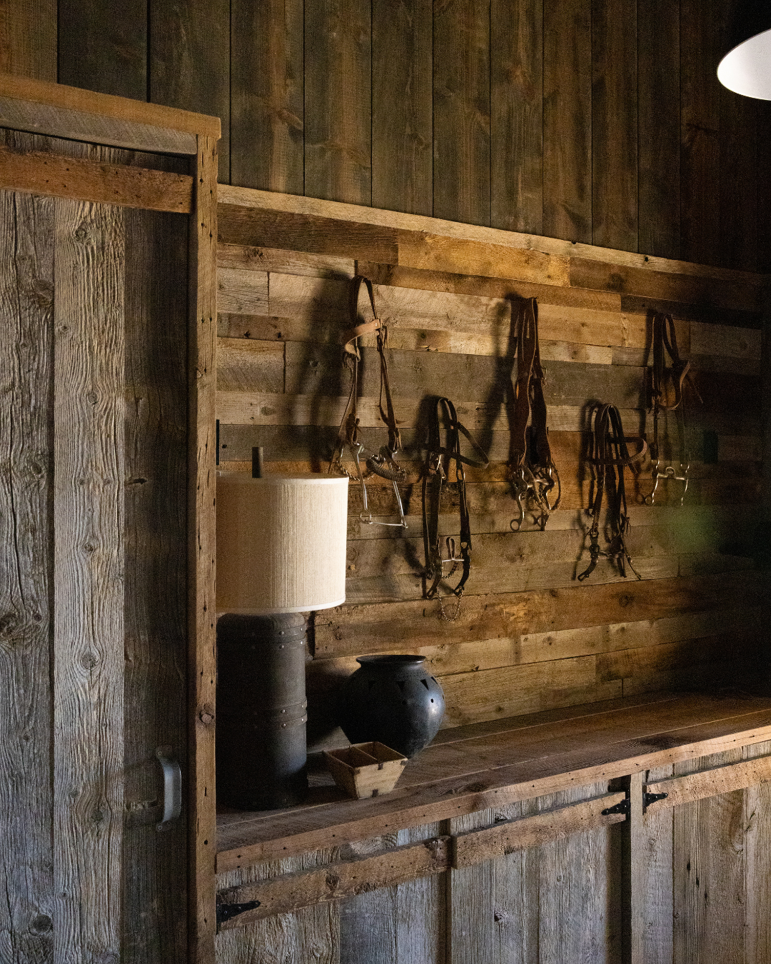 Riding bridles hanging on a rustic wooden wall in a barn with a lamp, a black vase, and a small basket on a wooden shelf.