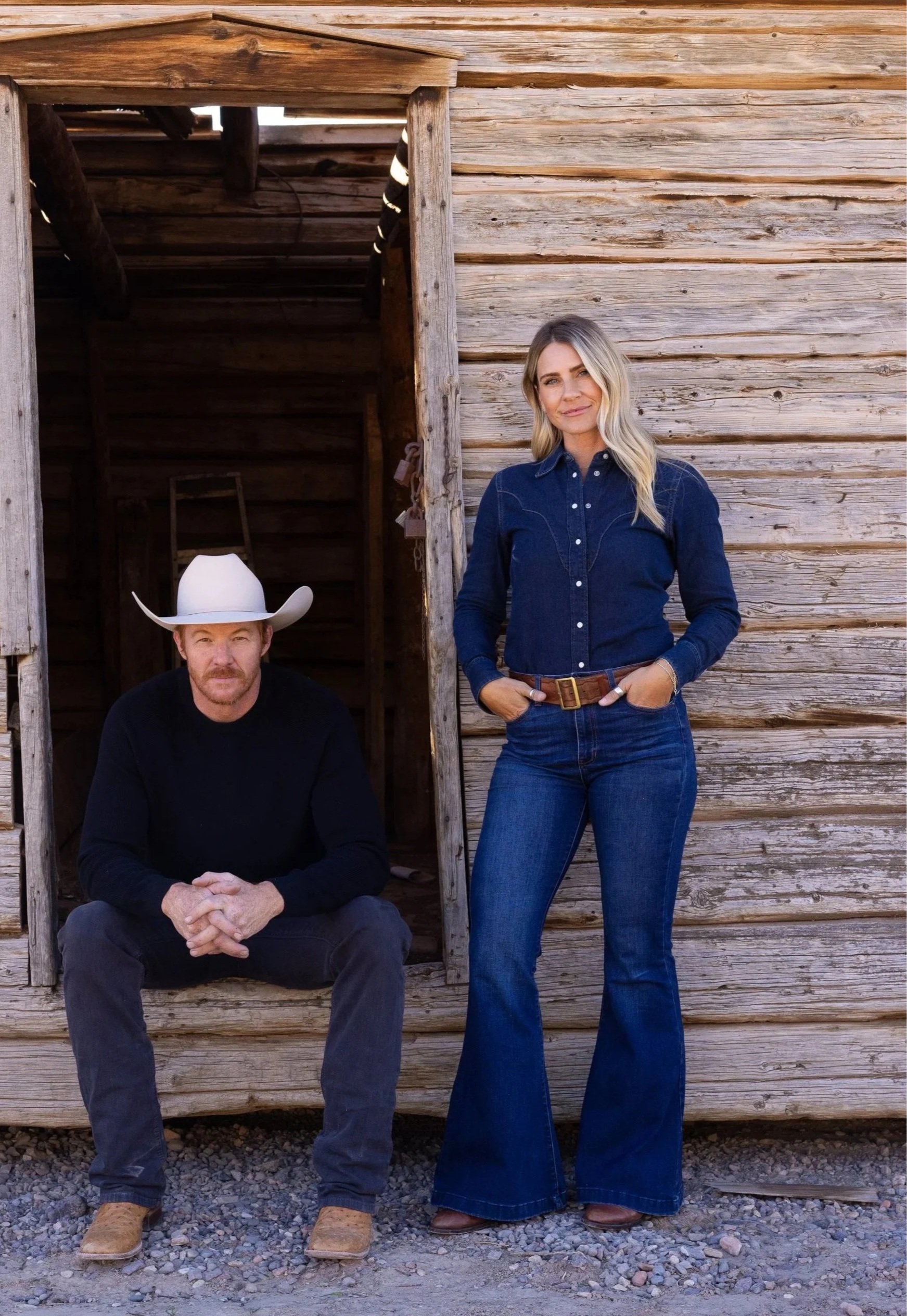 A woman standing next to a man sitting on the ground in front of a wooden building. The woman is wearing a blue denim shirt and jeans, while the man is wearing a black shirt, gray pants, and a white cowboy hat.