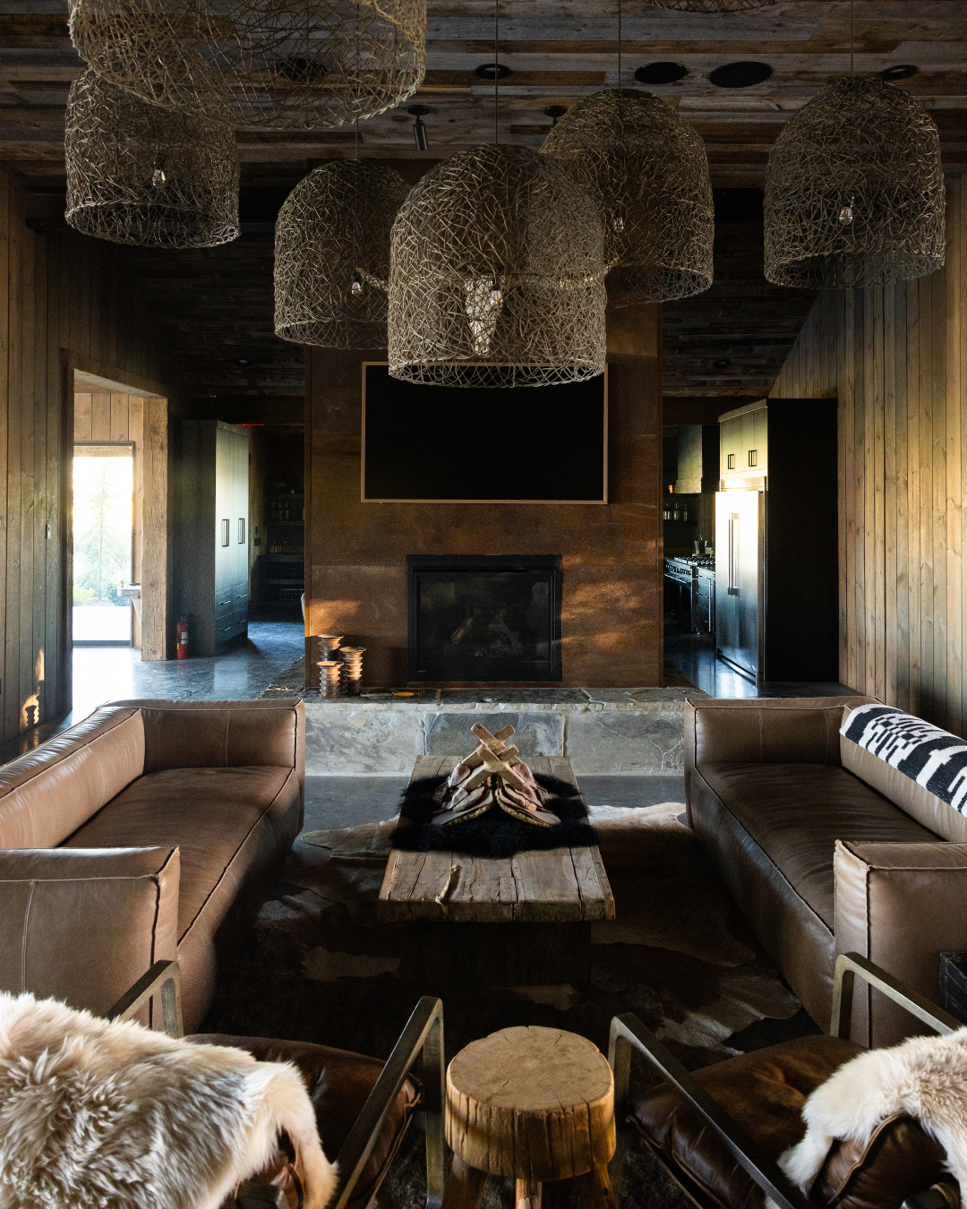 Living room with two leather sofas, a coffee table made from a wooden log, a fireplace, and a large flat-screen TV mounted above the fireplace. The room has wooden walls and ceiling, with woven pendant lights hanging from the ceiling. There are anima