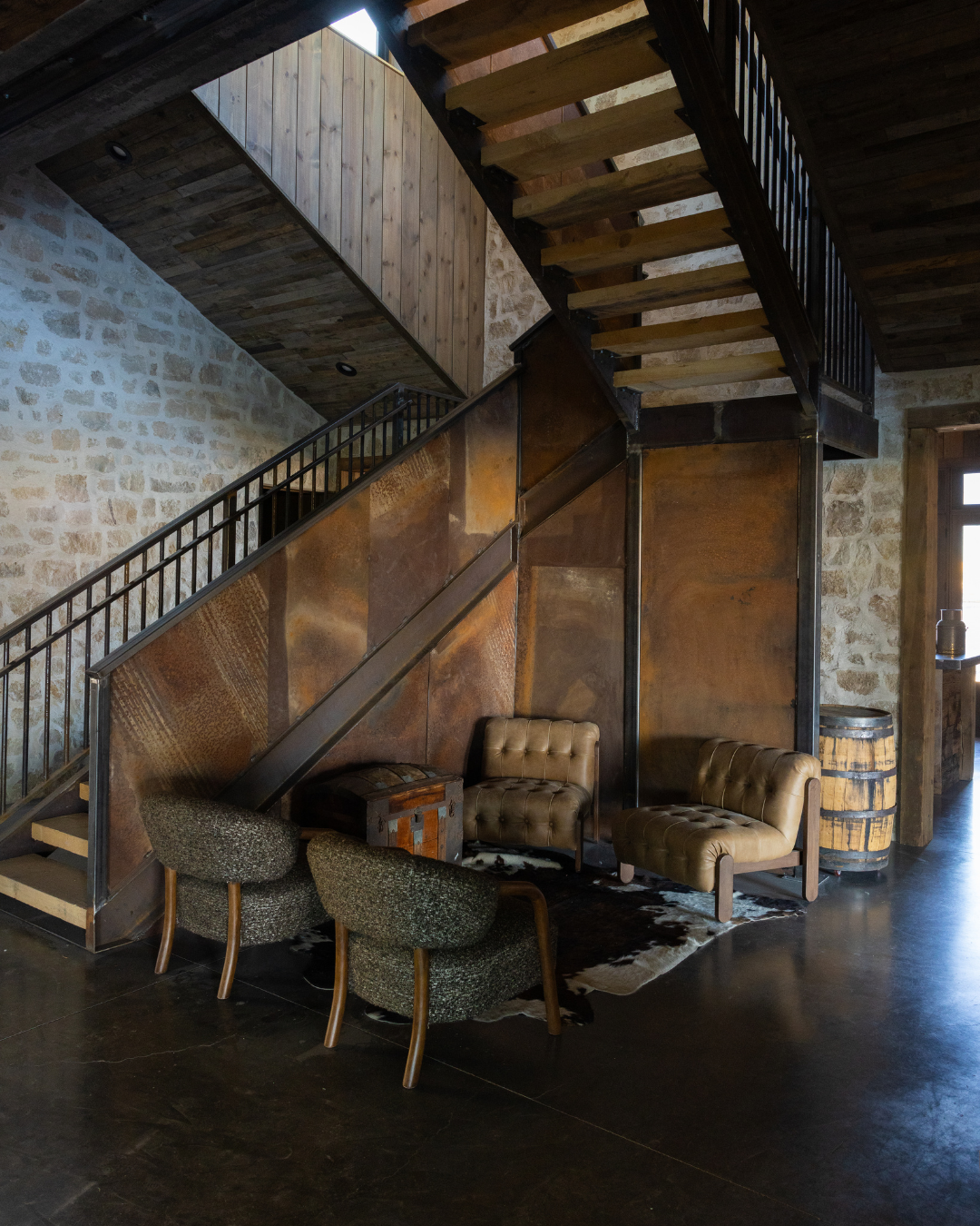 Interior of a rustic-style room with a staircase, three chairs, a barrel, and wooden walls.