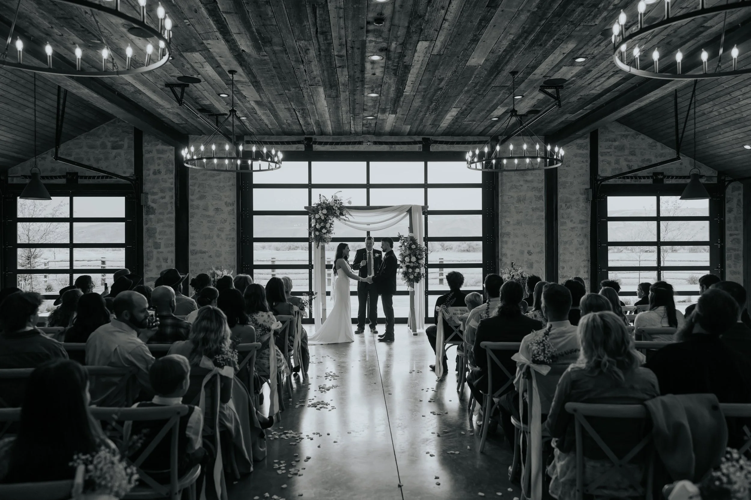 Indoor wedding ceremony with a bride and groom facing each other, officiant between them, surrounded by seated guests, large windows, rustic wooden ceiling, and chandeliers.