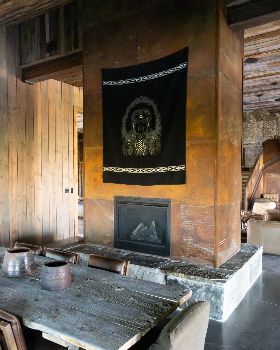 Cozy interior with a rustic wooden dining table, ceramic pots, a stone fireplace, and a black tapestry with an indigenous design hanging on a copper-colored wall.