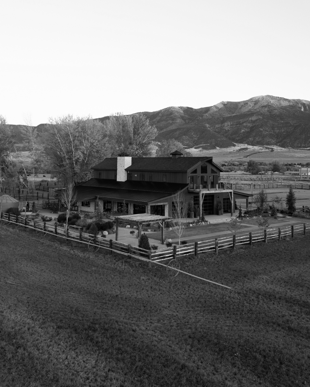 A black and white photograph of a large farmhouse with a chimney, surrounded by a fenced yard with trees, shrubs, and garden beds, with mountains in the background.