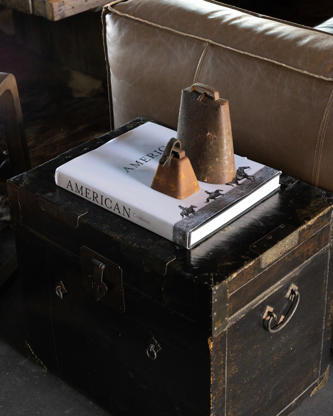 A black vintage trunk with metal accents, on which sits a large hardcover book titled 'AMERICAN' with images of cowboys, and two rusty iron weights.