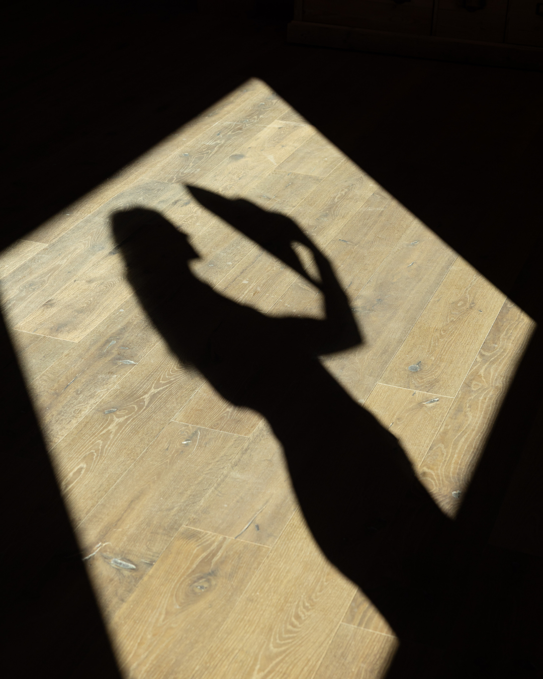 Silhouette of a person holding a cup, seen as a shadow on a wooden floor, with sunlight creating the shadow through a window.