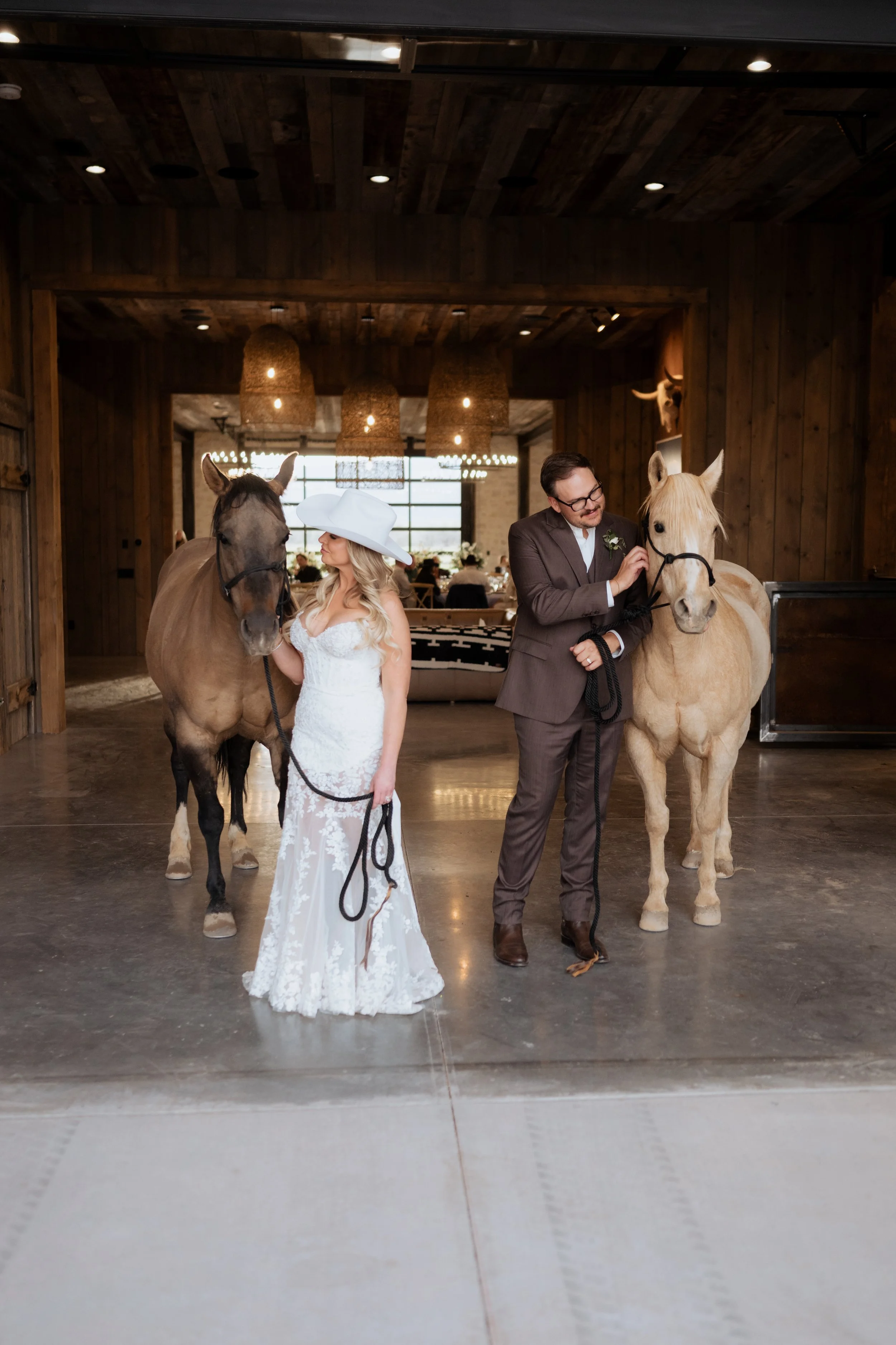 A bride and groom stand with two horses in a rustic wooden venue, with the bride in a white lace dress and wide-brimmed hat, and the groom in a brown suit, both holding the horses.