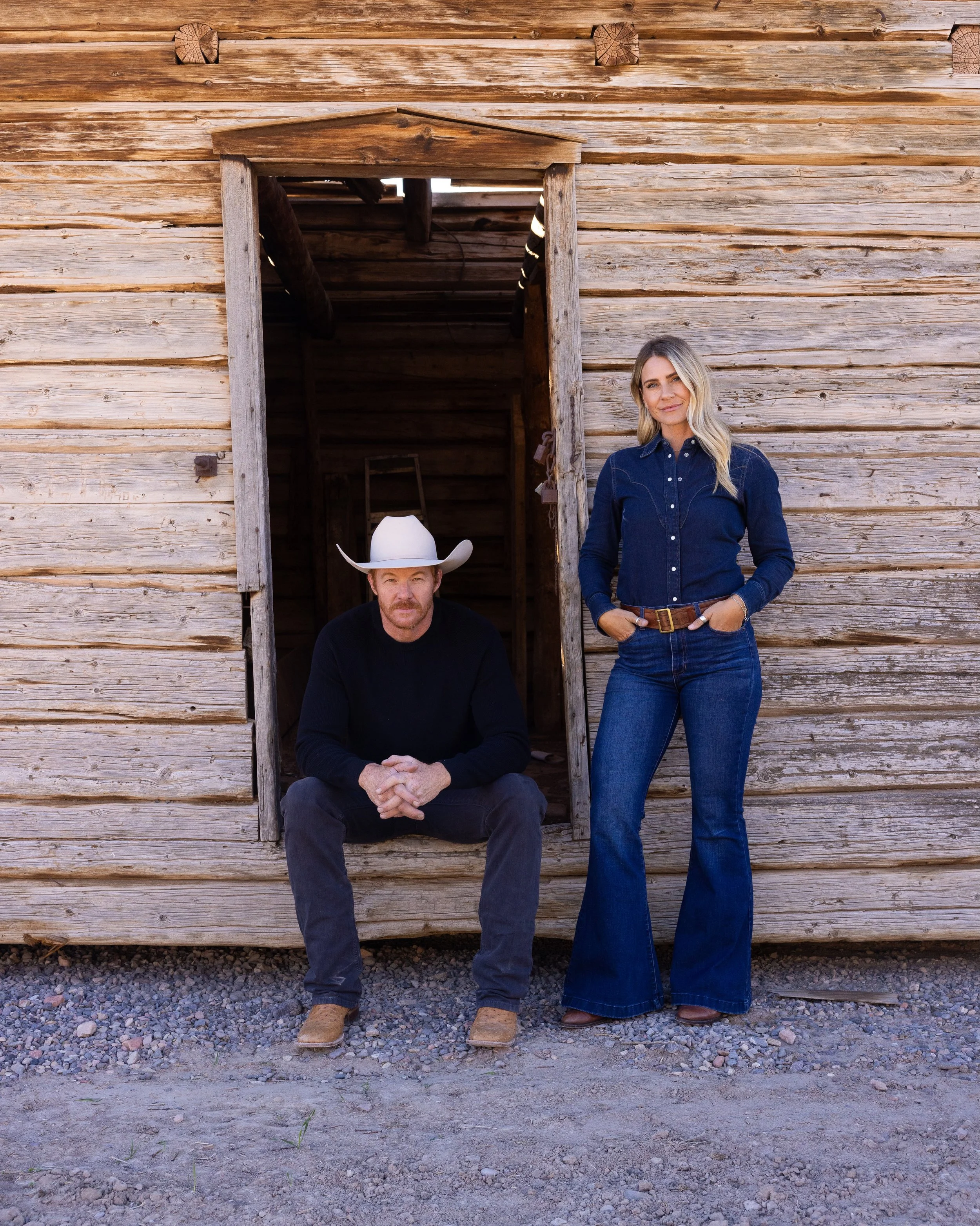 A man wearing a cowboy hat and a woman in a denim shirt posing in front of a weathered wooden building.