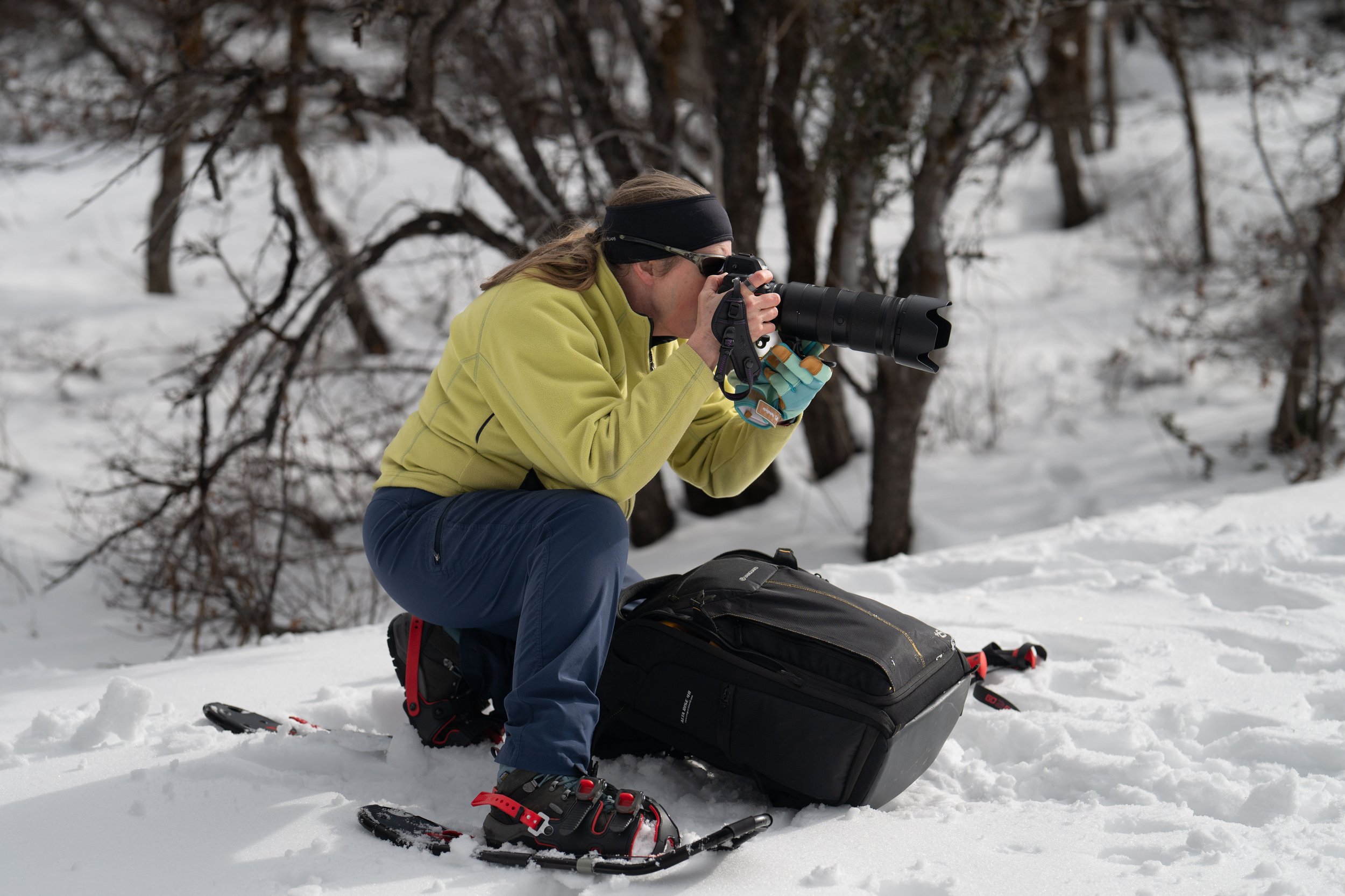 A woman wearing a yellow jacket, blue pants, and snowshoes kneeling in the snow, taking a photo with a camera equipped with a telephoto lens in Park City Utah. There is a black bag on the snow beside them and trees in the background.
