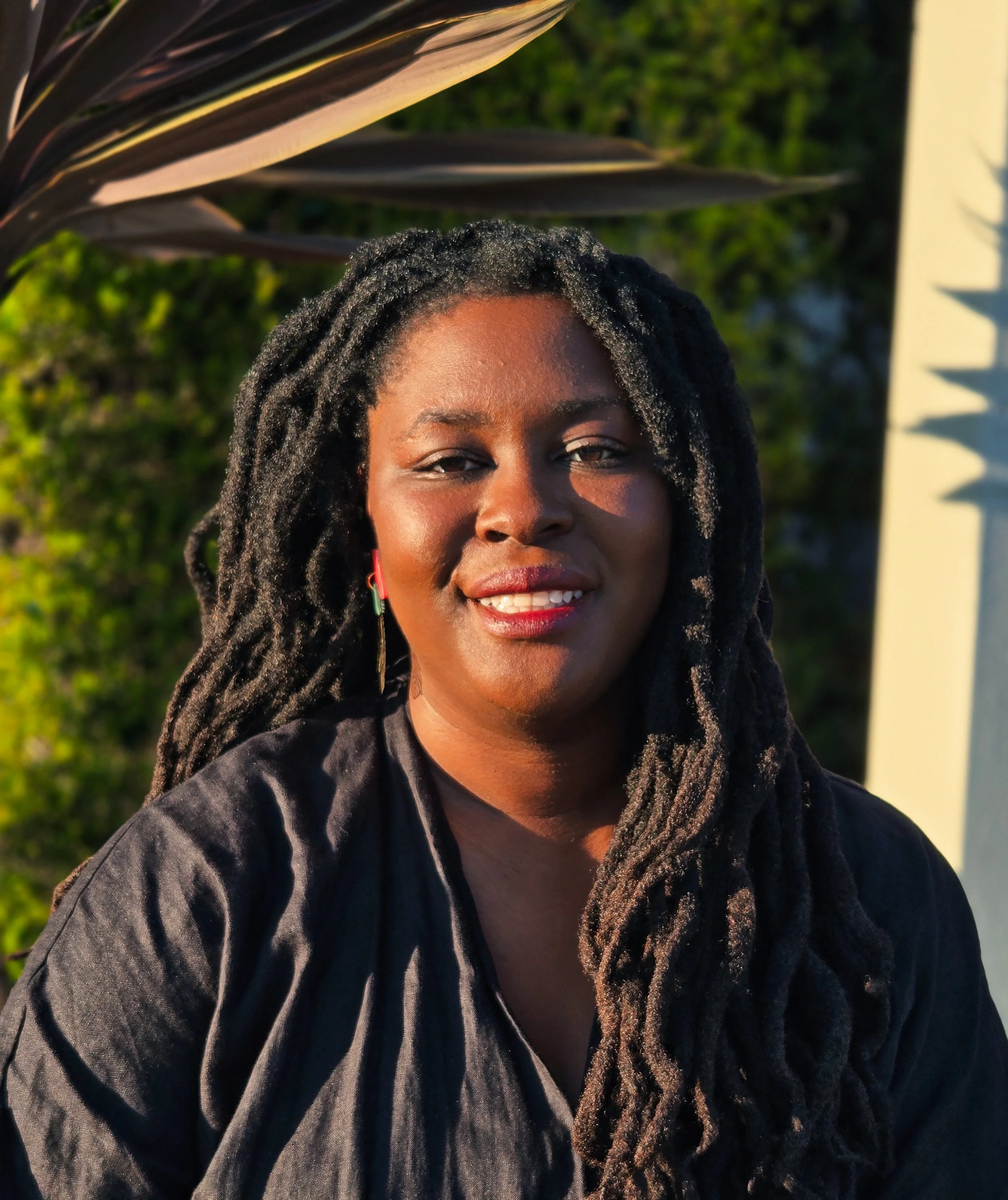 A smiling Black woman with long dreadlocks, wearing a black shirt and earrings, standing outdoors with green foliage and a sunlit background.