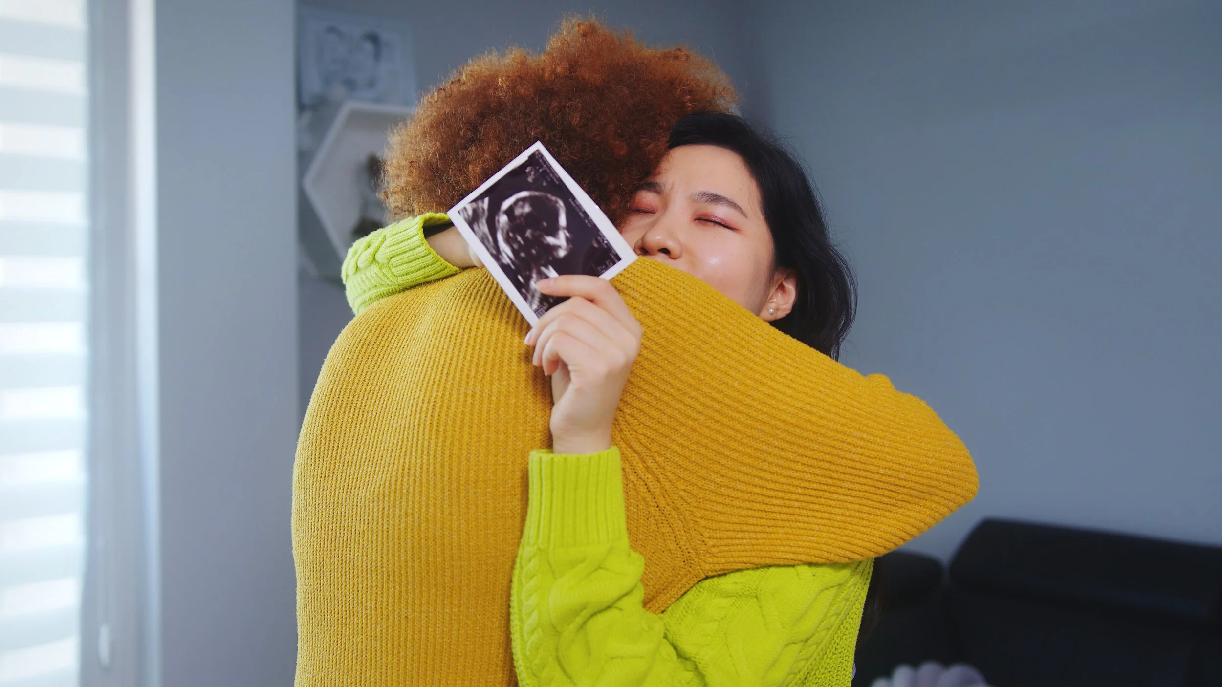 Two women hugging, one holding an ultrasound picture, in a warmly lit room.