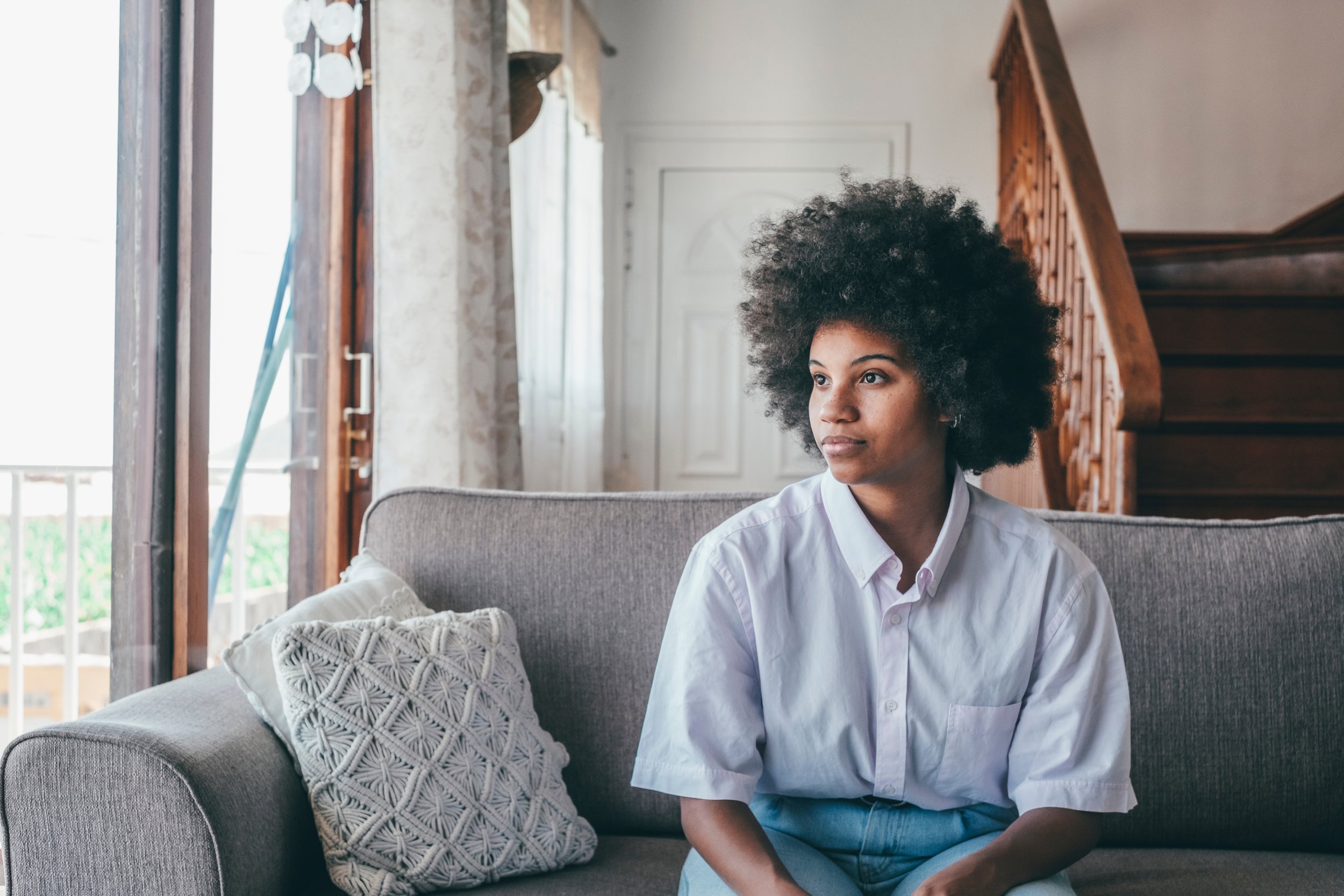A young woman with curly hair sitting on a gray sofa in a living room, looking thoughtfully to the side. The room has wooden stairs and a door in the background, with sunlight coming through an open window.