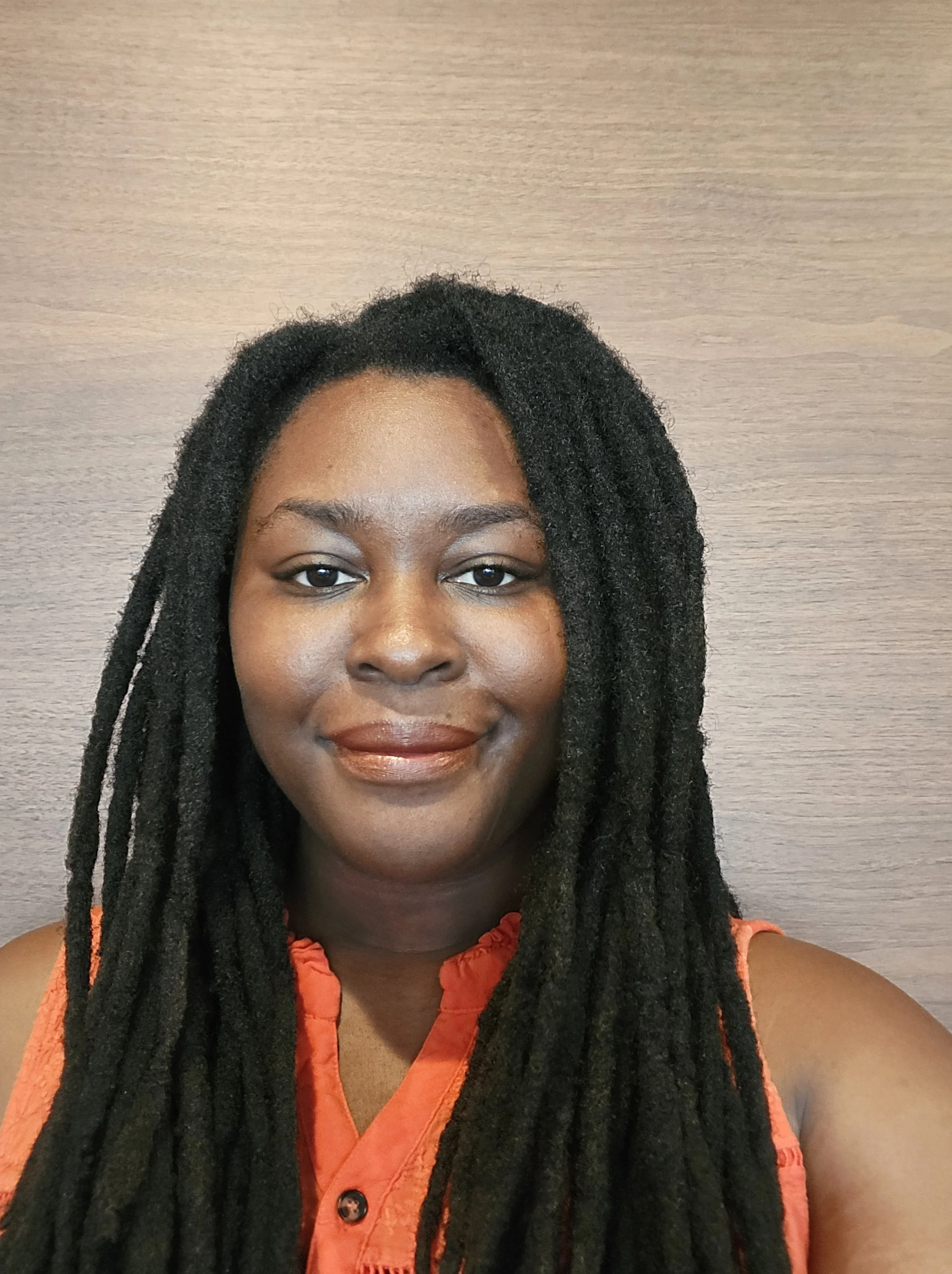 A woman with long black dreadlocks wearing an orange sleeveless top, standing in front of a wood-paneled wall.